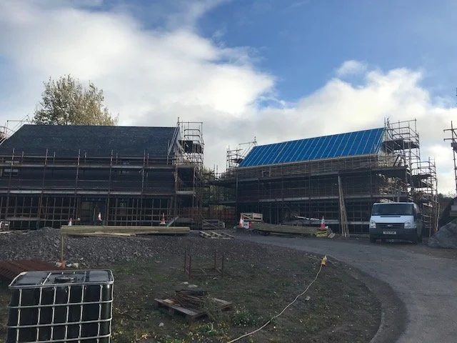 Two houses under construction with scaffolding, one with a black roof and the other with a blue roof, on a partly cloudy day with construction materials nearby.