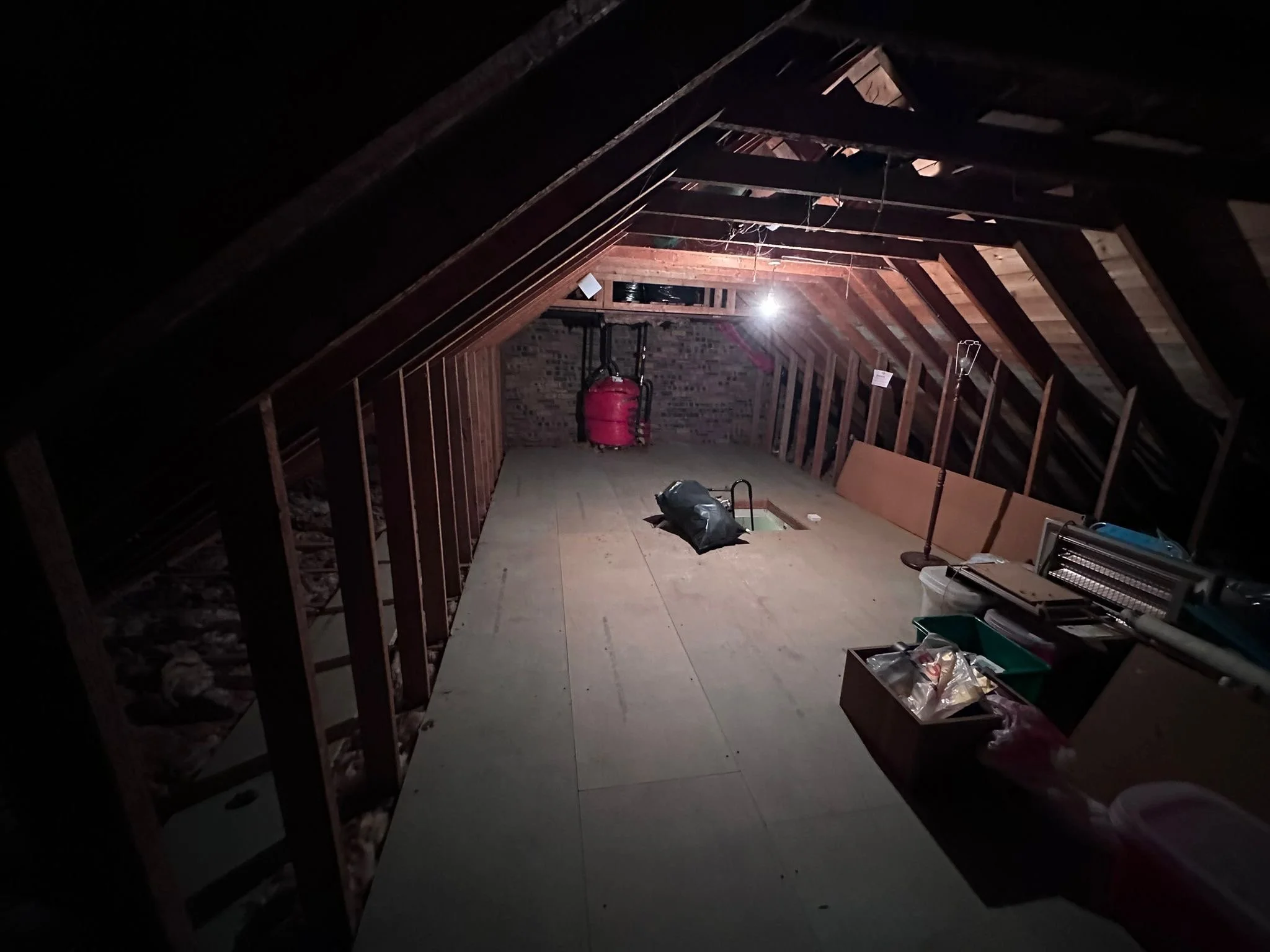 Attic with exposed wooden beams, lighting coming from a small bright light, storage items including a pink vacuum cleaner and plastic bags, and various boxes and supplies.