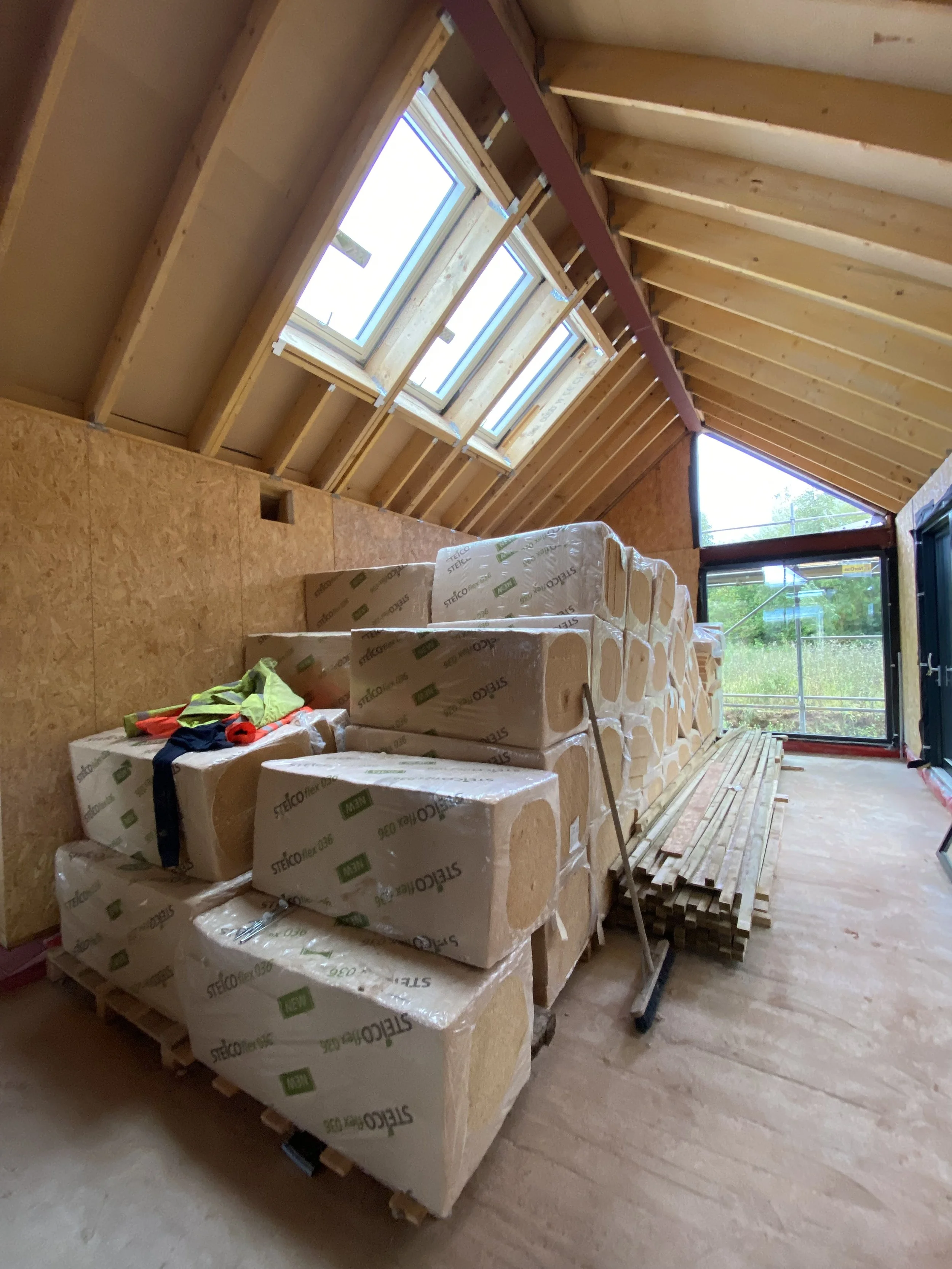 An indoor construction site with wall insulation stacked on wooden pallets, window frames installed in the ceiling, and a stack of wooden planks on the floor.