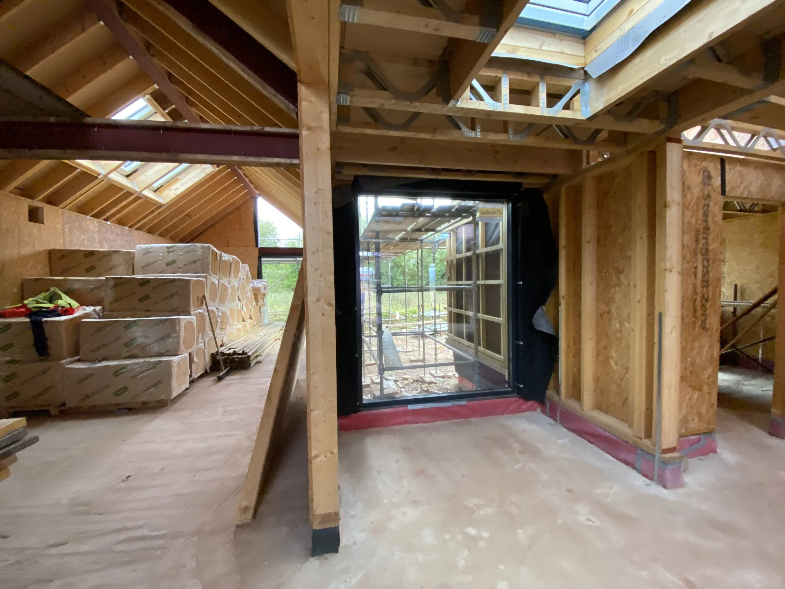Interior of a house under construction with exposed wooden framing, a newly installed window, and boxed electrical wiring and insulation materials.