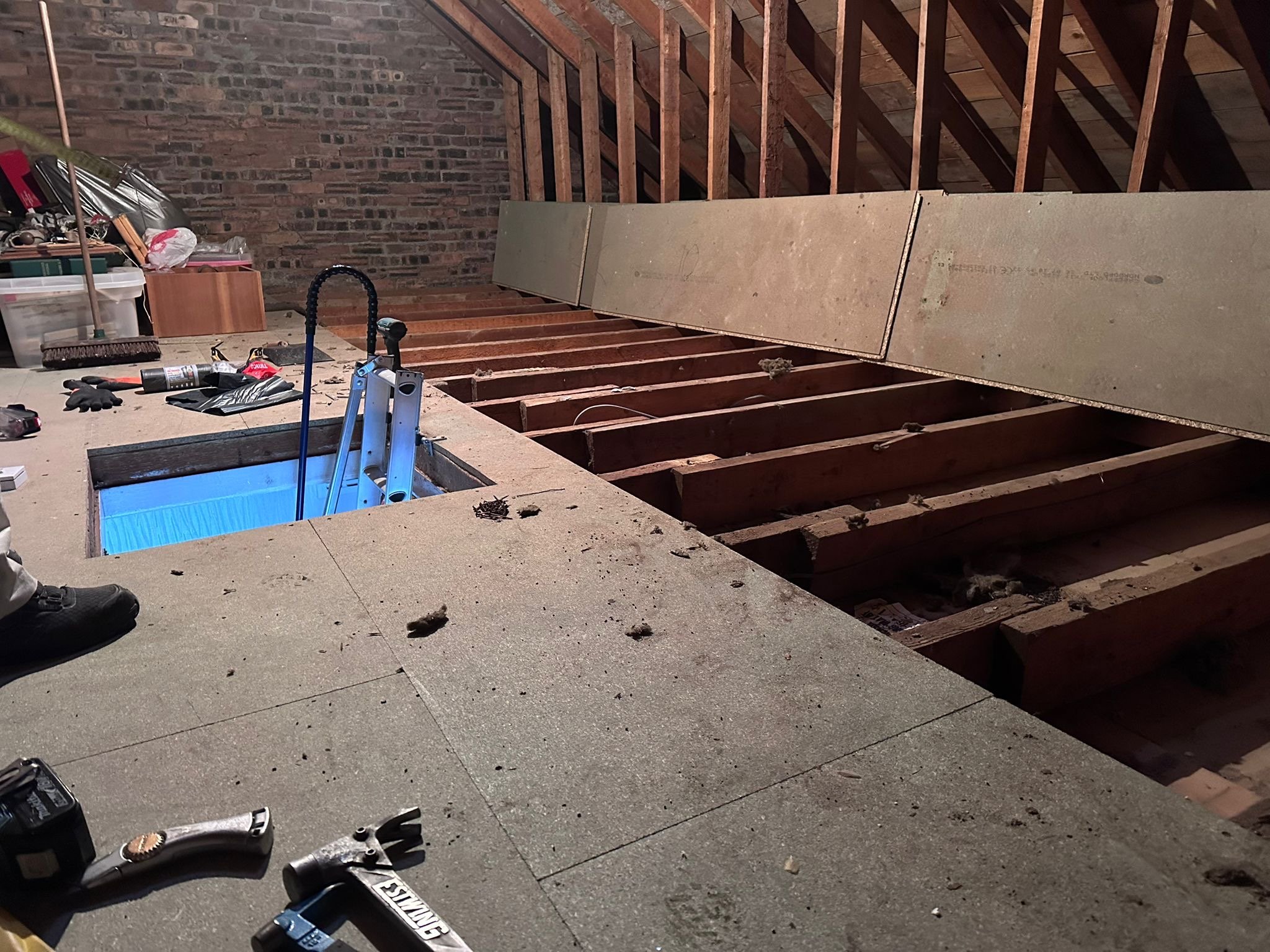 Attic space under renovation with exposed wooden floor joists and partial wall insulation. Tools, gloves, and construction materials are scattered around, with a brick wall visible in the background.