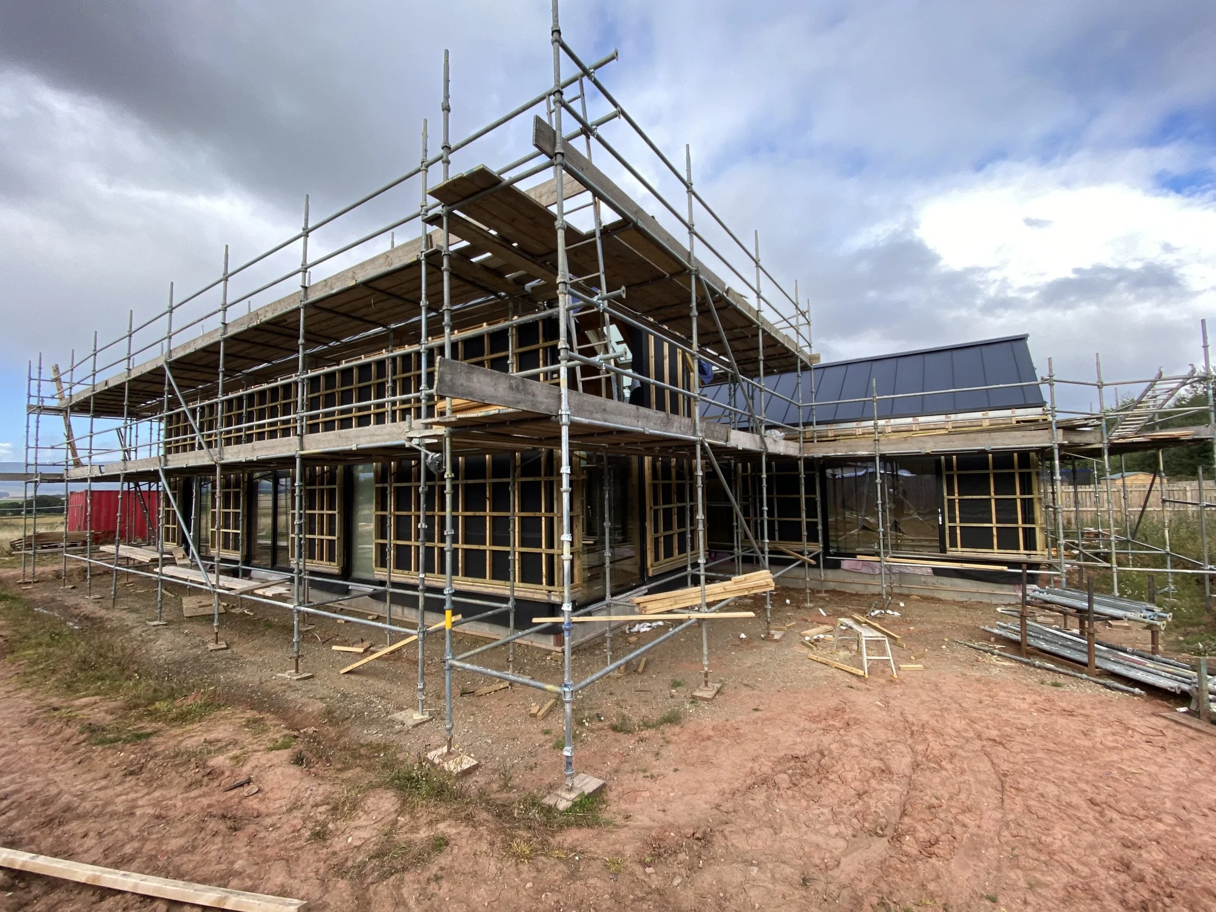 Construction site with a partially built house surrounded by metal scaffolding, on a cloudy day with a dirt ground in the foreground.
