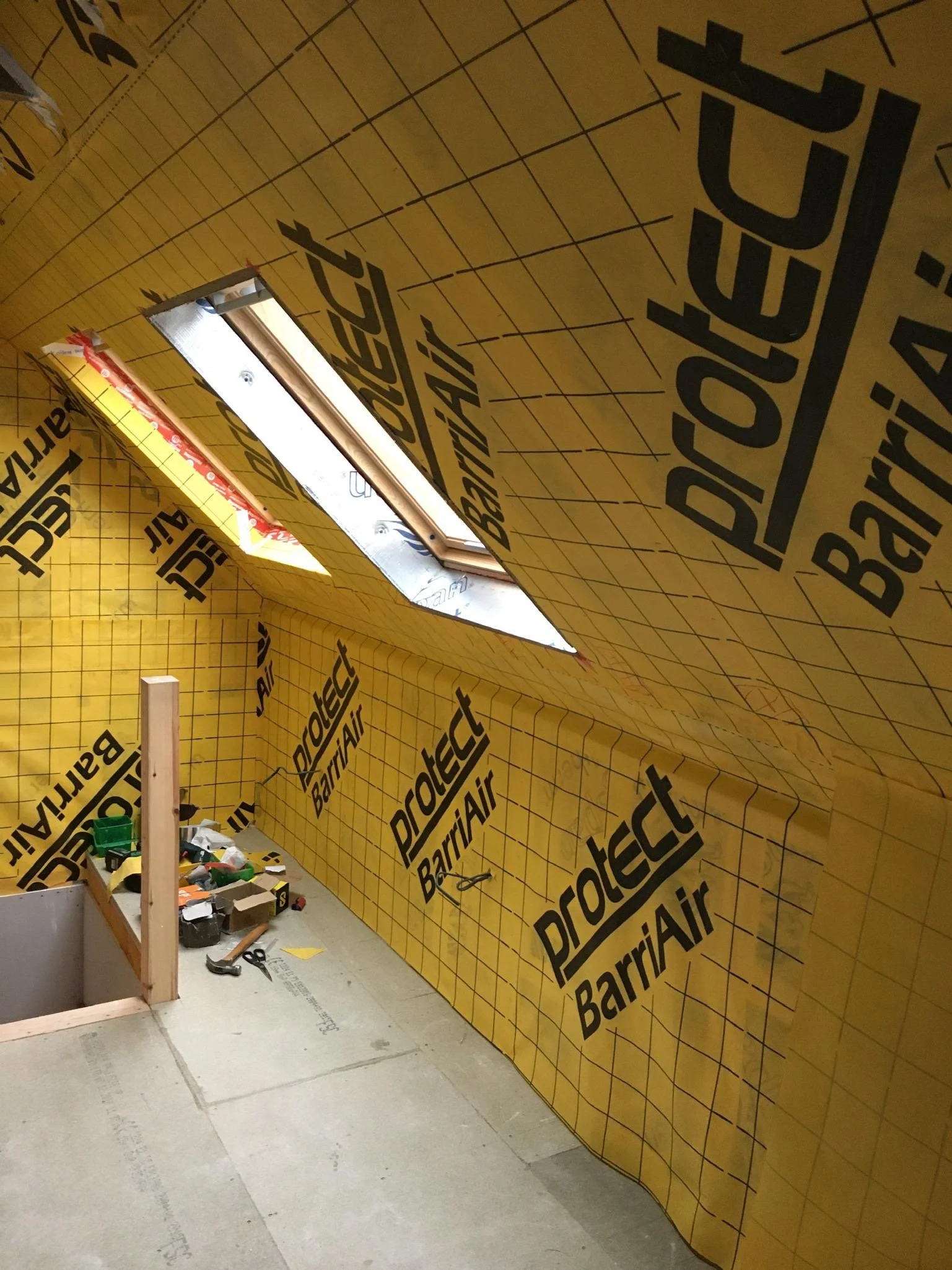 Interior of a building under construction or renovation with yellow Barricade barrier wrap on walls, a window opening, and construction tools and materials on the floor.