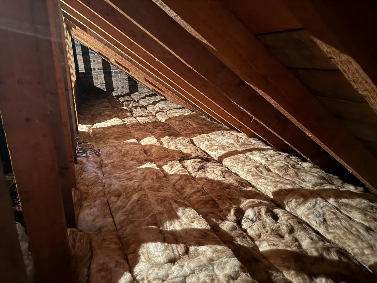 Attic space with exposed wooden beams and insulation material on the floor, a brick wall at the far end, and electrical wires.