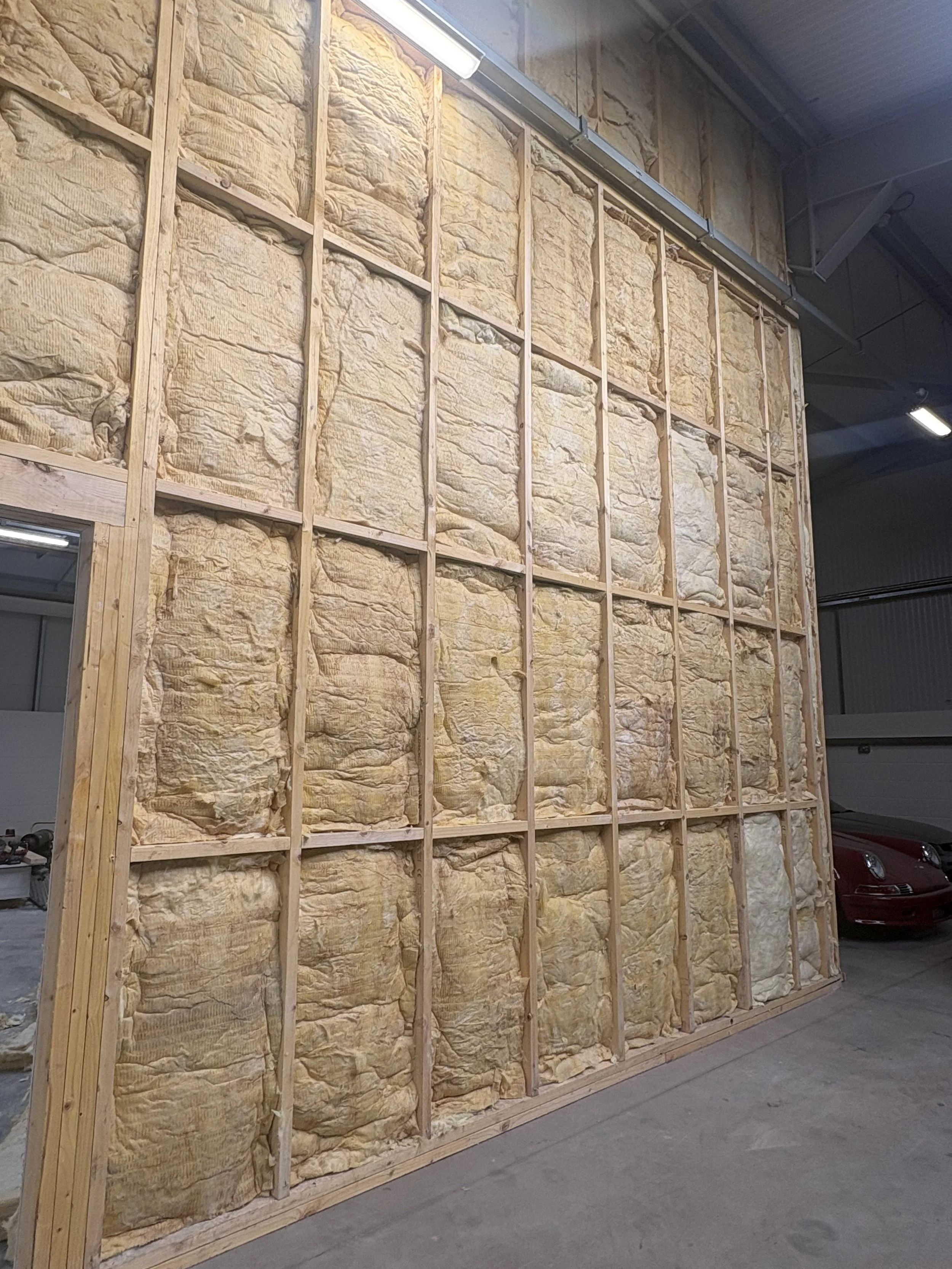 Wooden framing of a wall with insulation material installed between the studs in an indoor construction site, with part of a garage and a red car visible on the right.