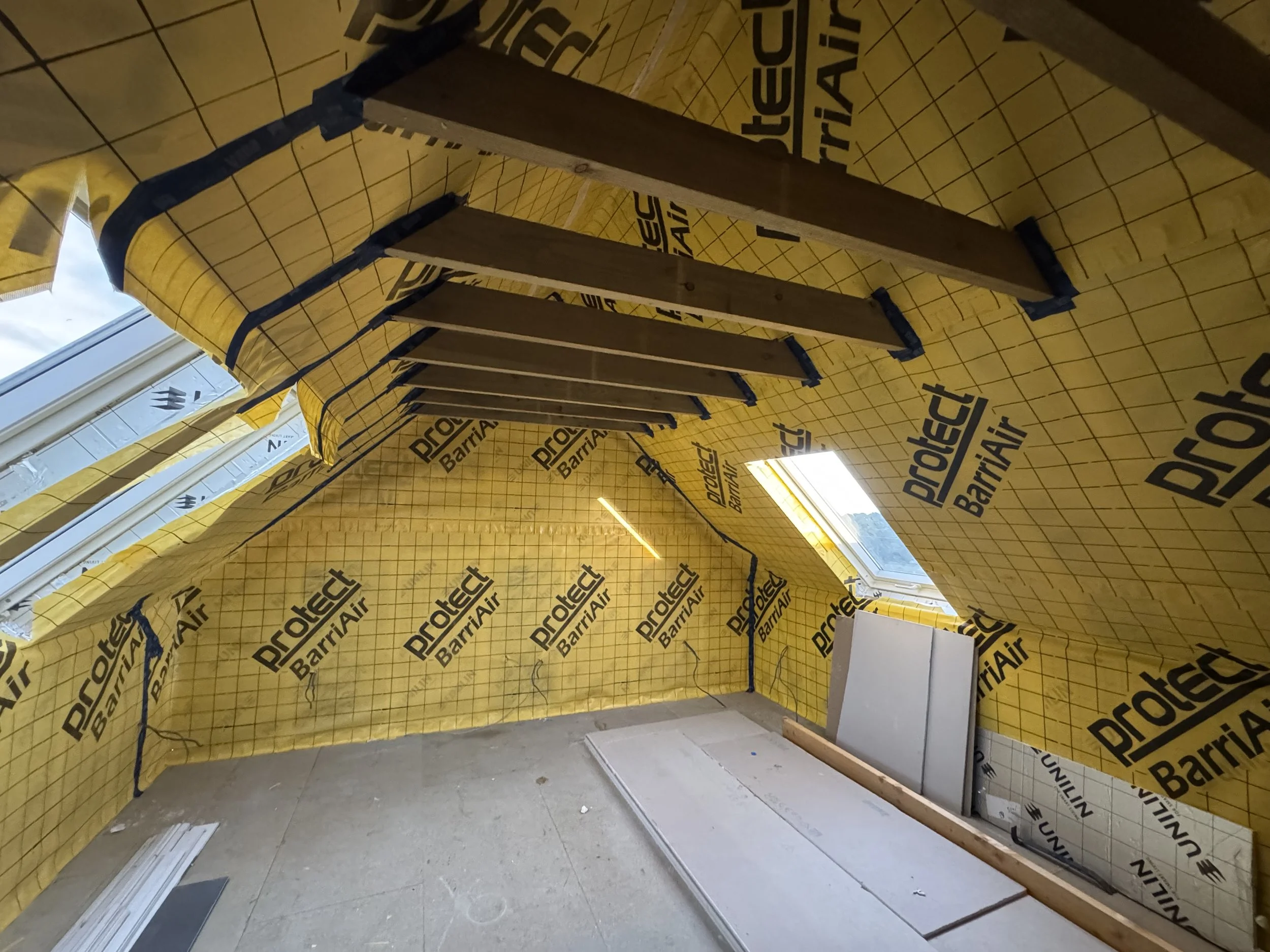 Interior of a room under construction with yellow insulation material labeled 'ProtEnd Barrie' on walls and ceiling, with wooden framing, window, and some electrical wiring visible.