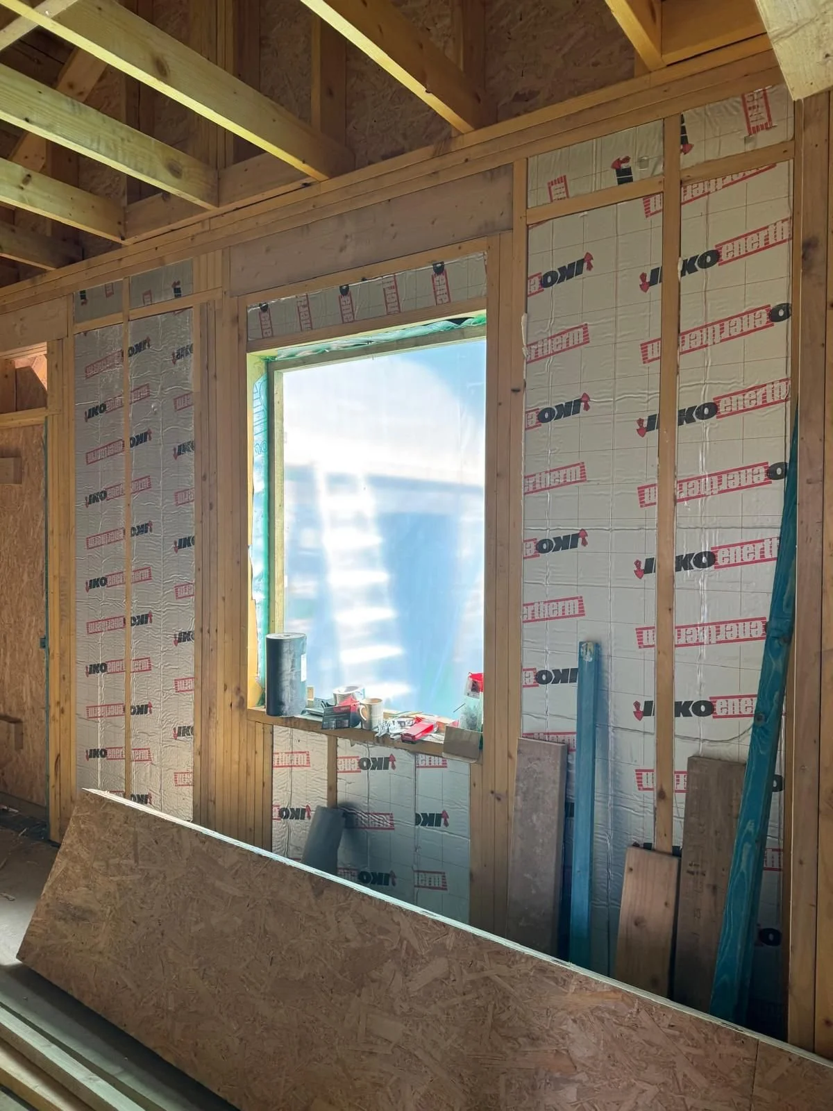 Construction site of a wooden framed building with insulation panels installed on the walls, a window opening covered with plastic, construction tools on the window sill, and wooden planks leaning against the wall.