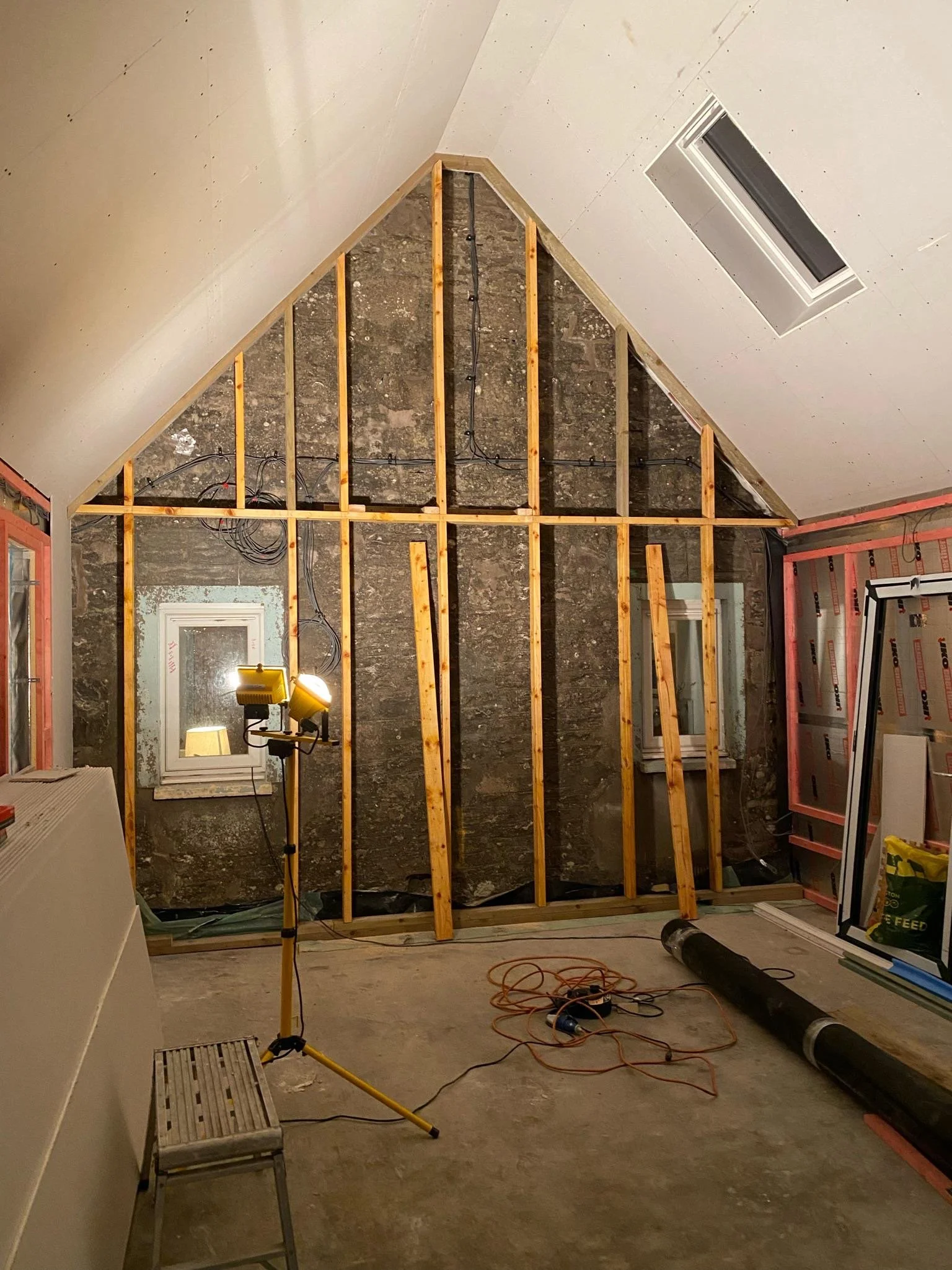 A room under construction with exposed wooden framing and insulation, a window, and construction tools and materials scattered on the floor.
