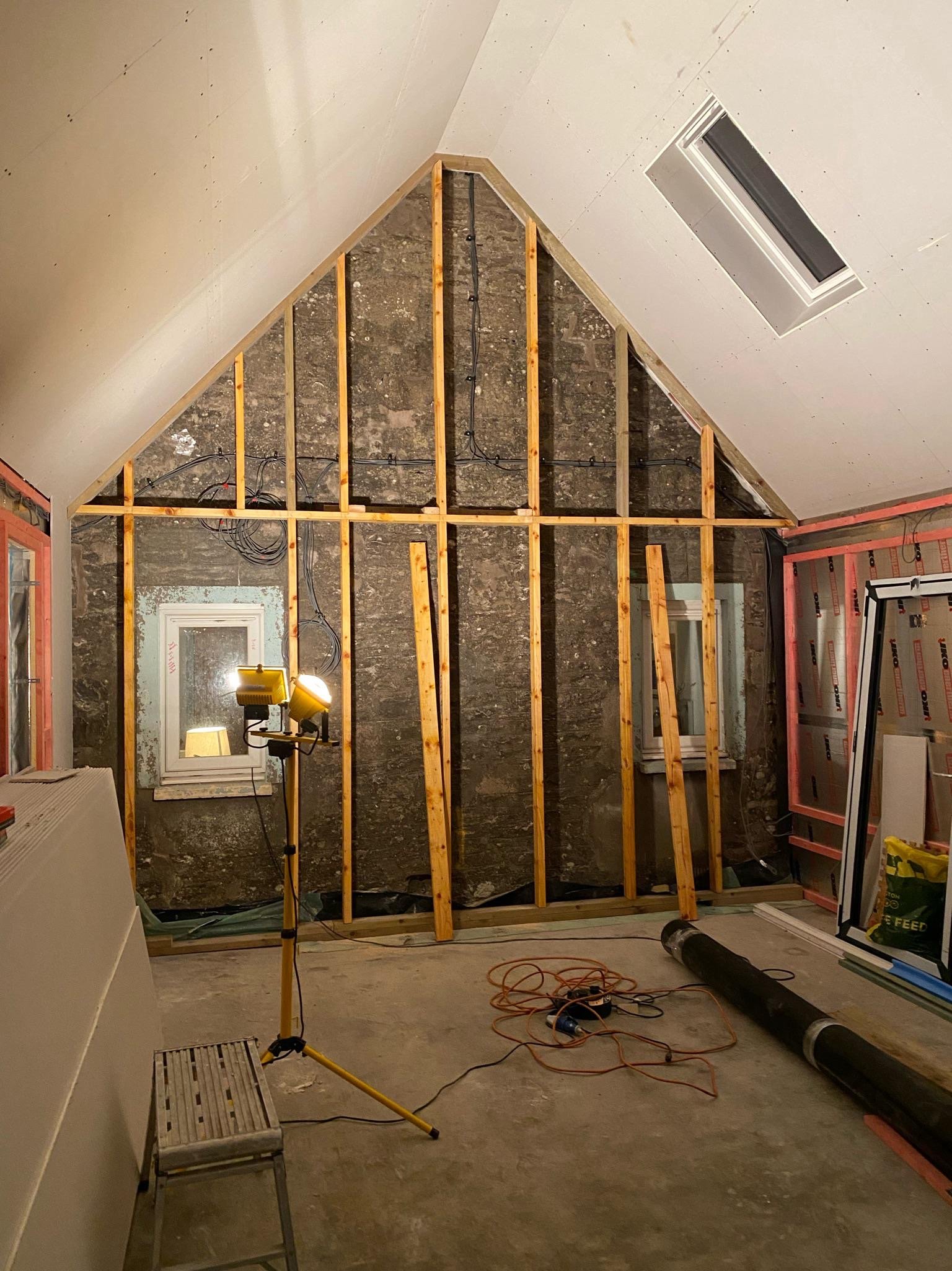 Interior of a room under renovation with exposed wall studs, electrical wiring, construction tools, and an angled ceiling with a skylight.