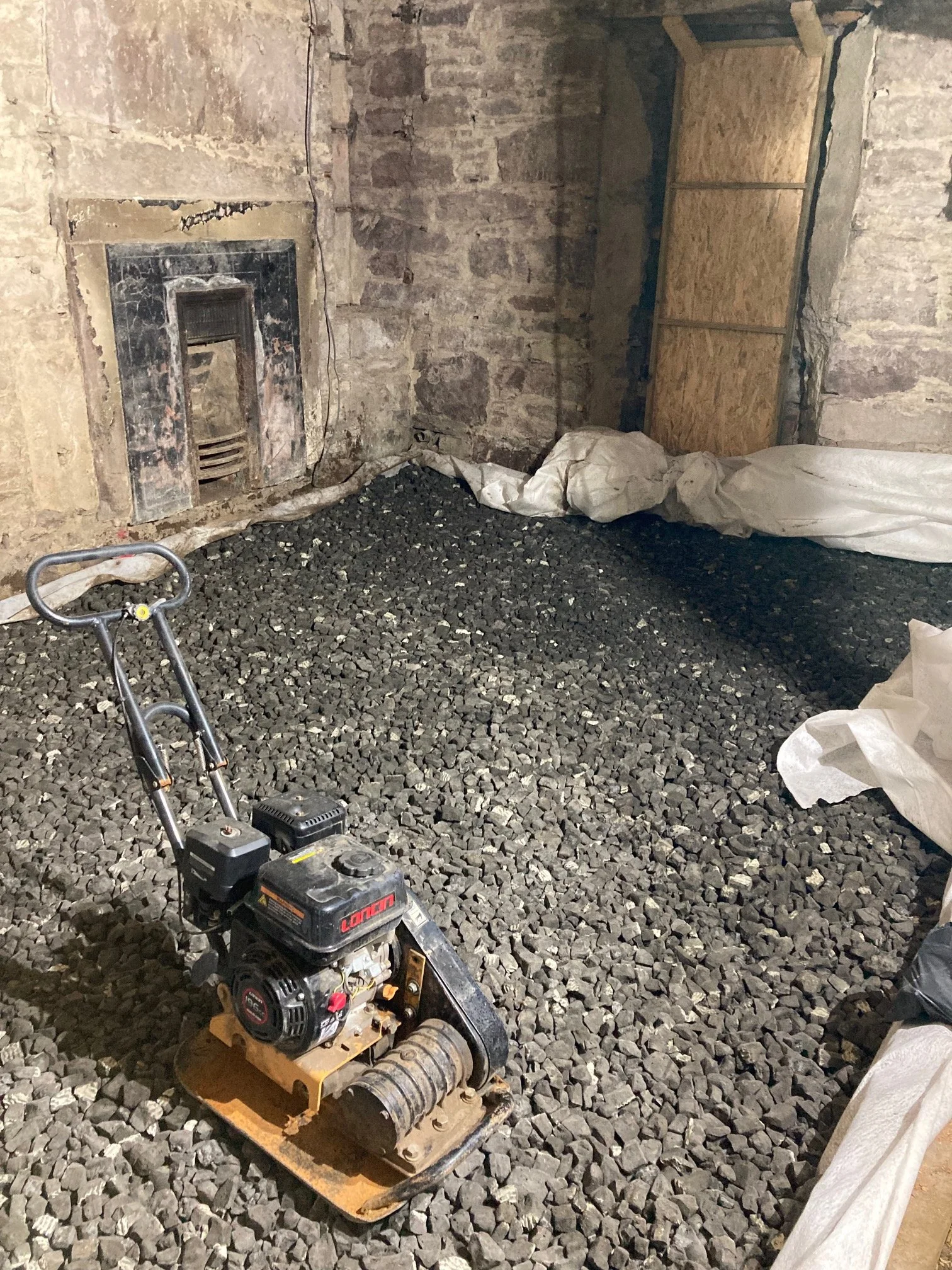 A basement under construction with a stone wall and a fireplace. The floor is covered with gravel. A small gas-powered compactor is in the foreground. There is a large piece of white fabric or plastic on the floor to the right and a wooden panel on the wall on the right side.