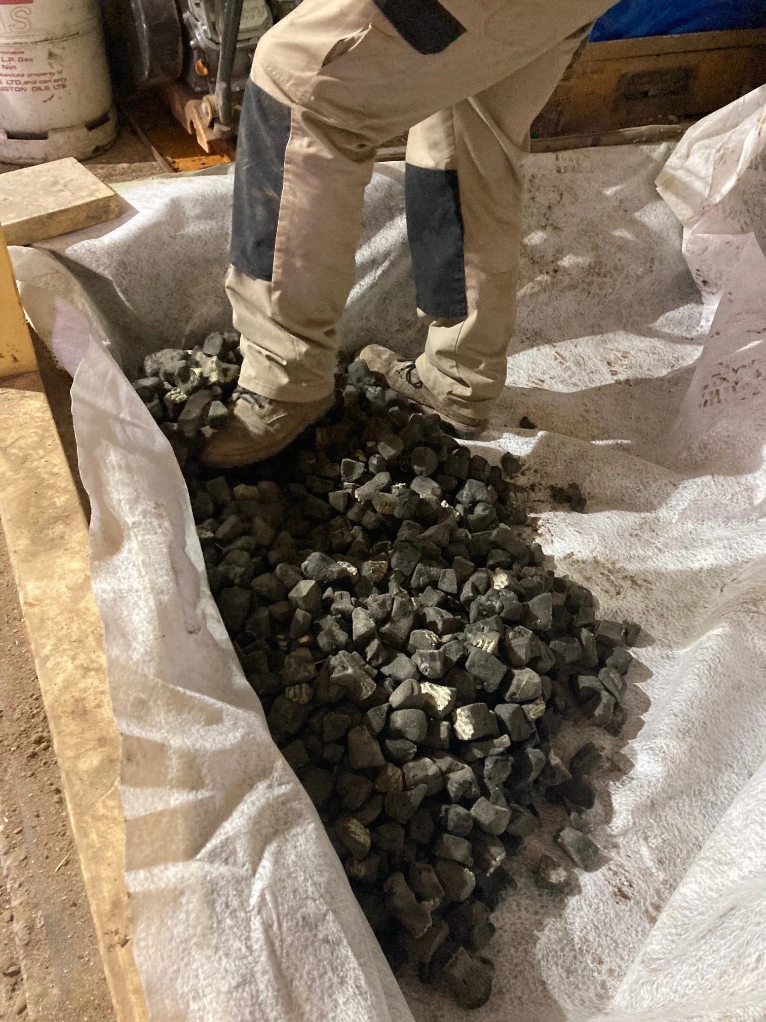 Person wearing beige work pants and shoes stands next to a large white bag filled with small, dark stones inside a workshop or garage.