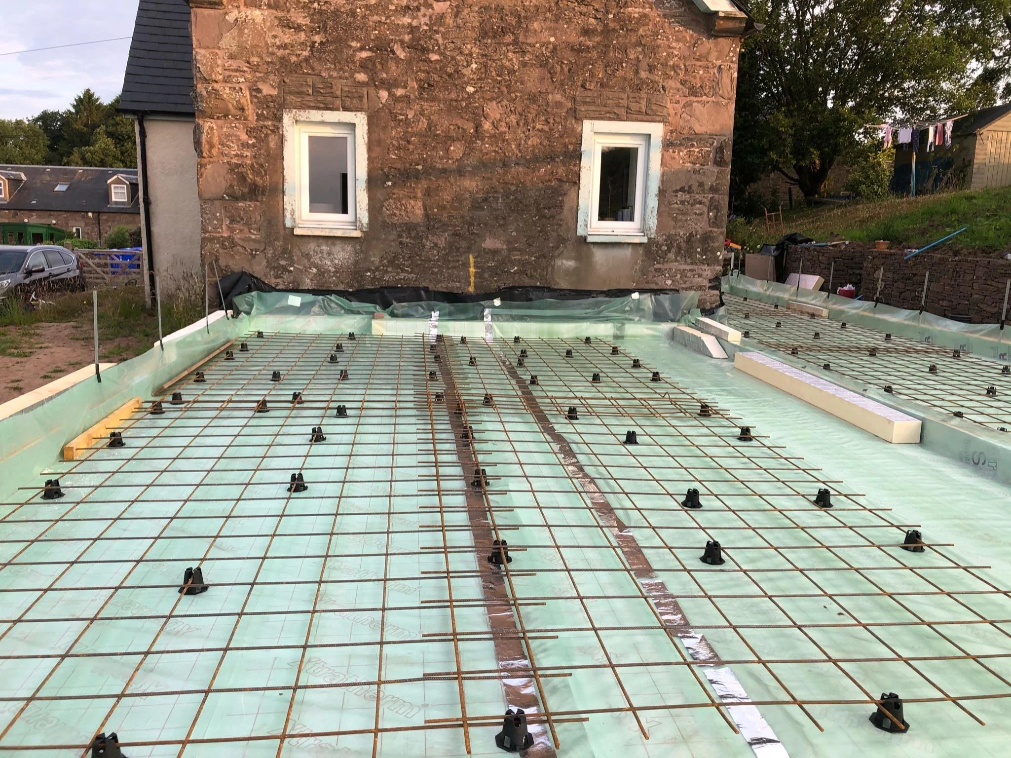 Construction site for a building foundation with rebar grid, foundations, and insulation, in front of a stone house with two small windows, and a yard with trees, a clothesline, and other houses in the background.