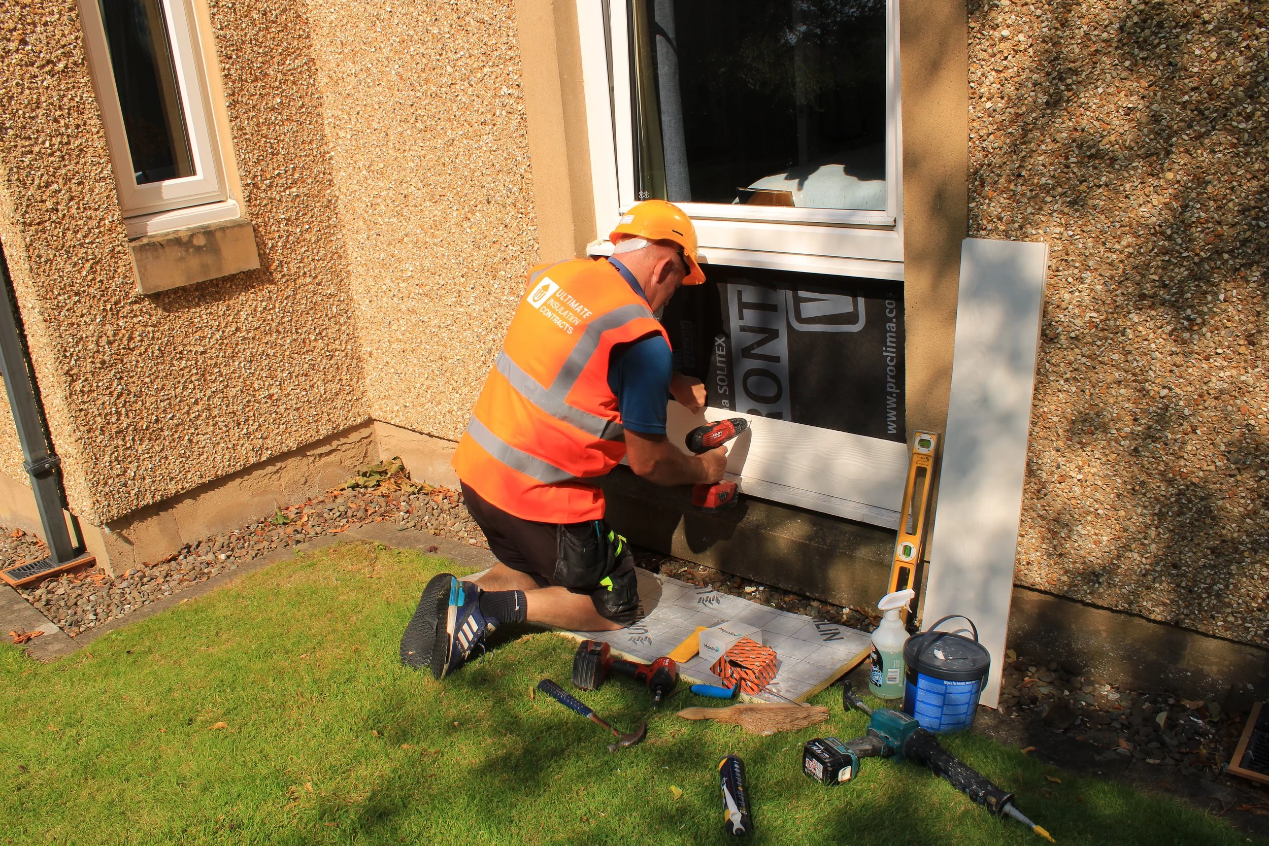 A construction worker kneels on the ground installing a window frame on a building exterior. He wears a yellow safety helmet and an orange safety vest with gray reflective stripes. Various tools and supplies are scattered around him, including a cord