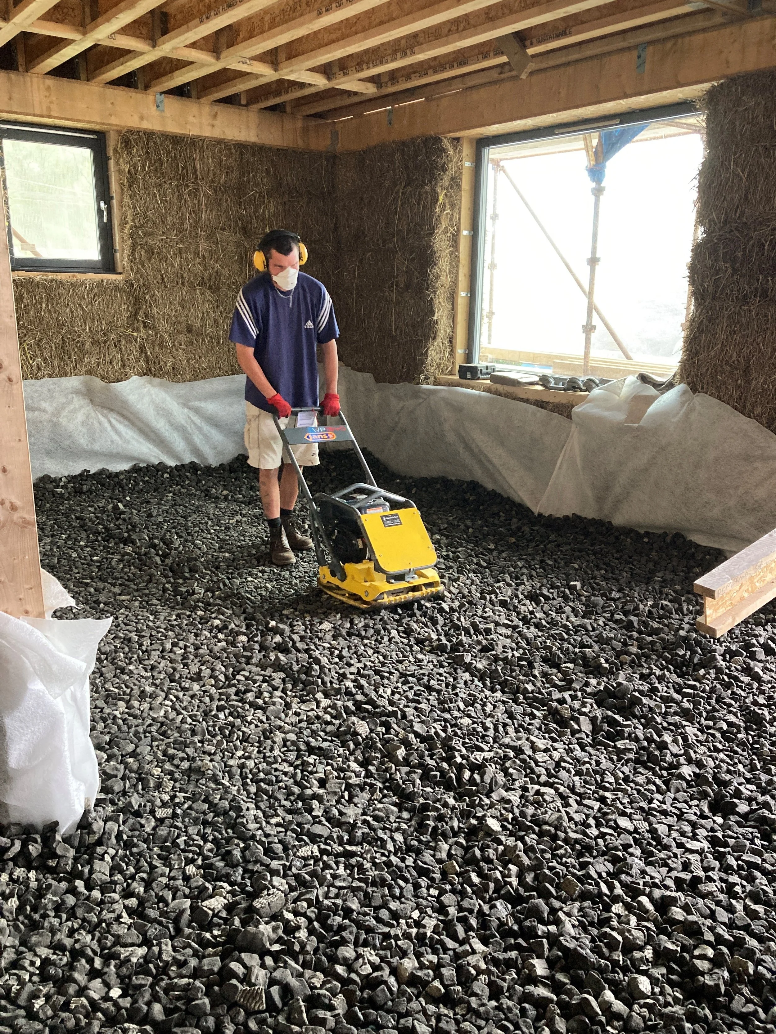 A person operating a compactor machine inside a partially constructed building, with wood framing and insulation materials visible, and a large window letting in natural light.