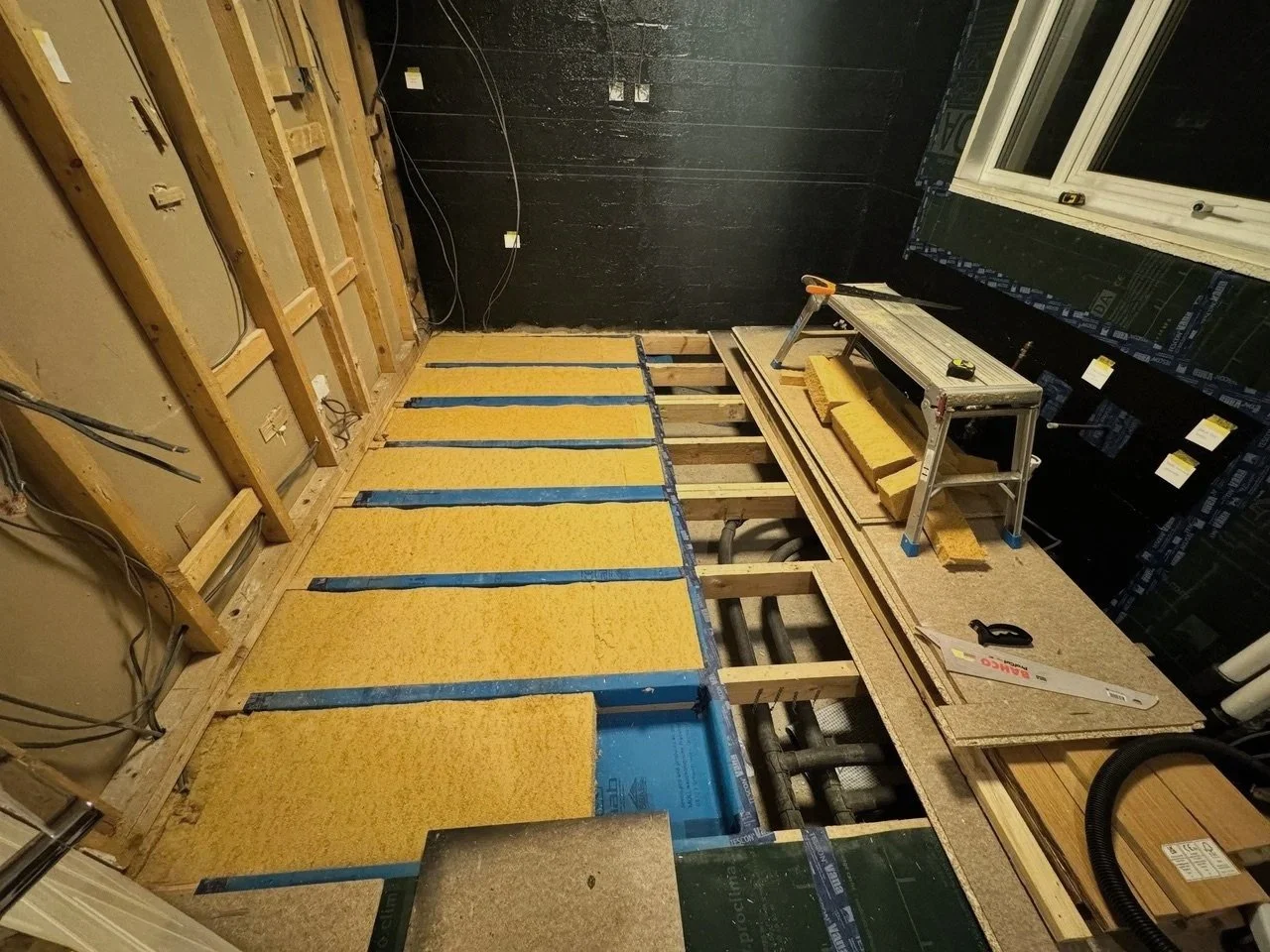 Room under construction with exposed wooden framing, yellow insulation material, black drywall, a worktable with construction tools, and a window.