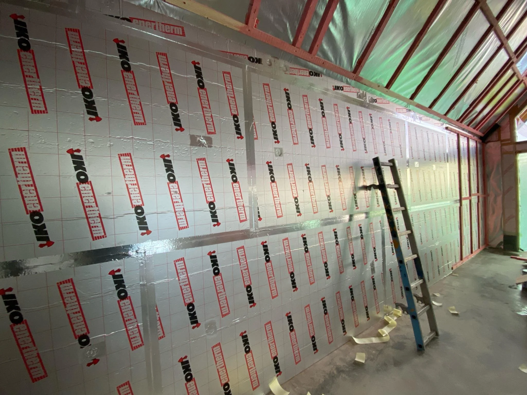 Interior view of a building under construction with insulated wall panels, wooden framing, and a ladder.