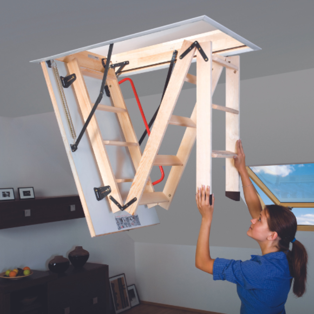 A person installing a wooden attic ladder through a ceiling hatch in a room with sloped ceiling and a window.
