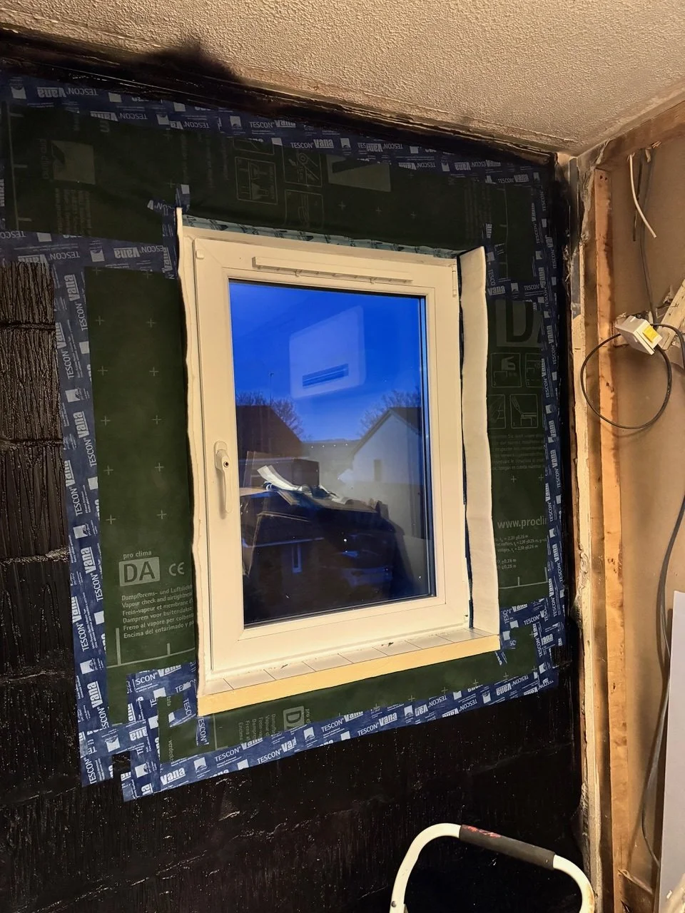 A window installed in a wall with black waterproofing material and green insulation sheets around it, in a room under construction.