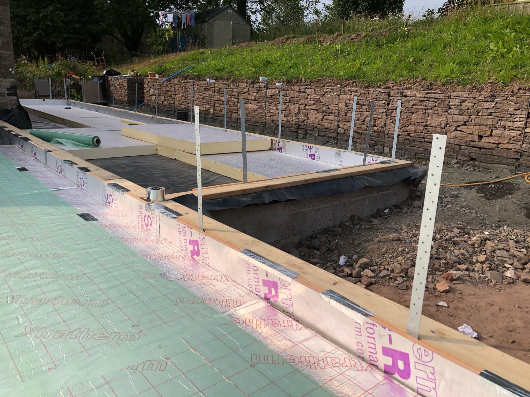 Construction site with foundation being prepared, insulation and barriers in place, backyard with green grass, stone retaining wall, trees, shed, and laundry hanging to dry in the background.