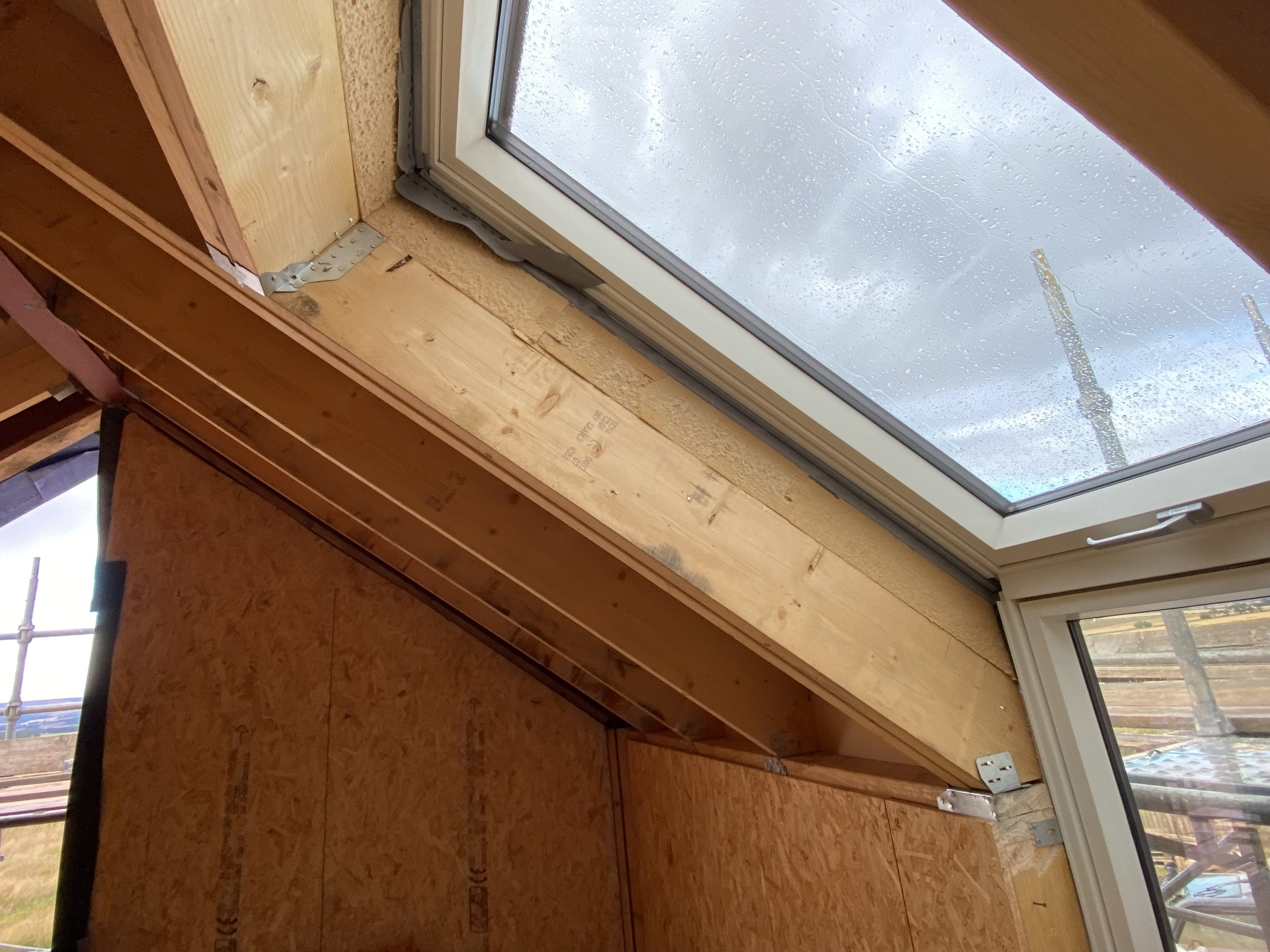 An interior view of a room showing partly unfinished walls with wood paneling and an angled ceiling with a large window. The window has sliding panes with water droplets on the glass, and outside, cloudy sky and some structural elements are visible. Scaffolding is partially visible outside the window.