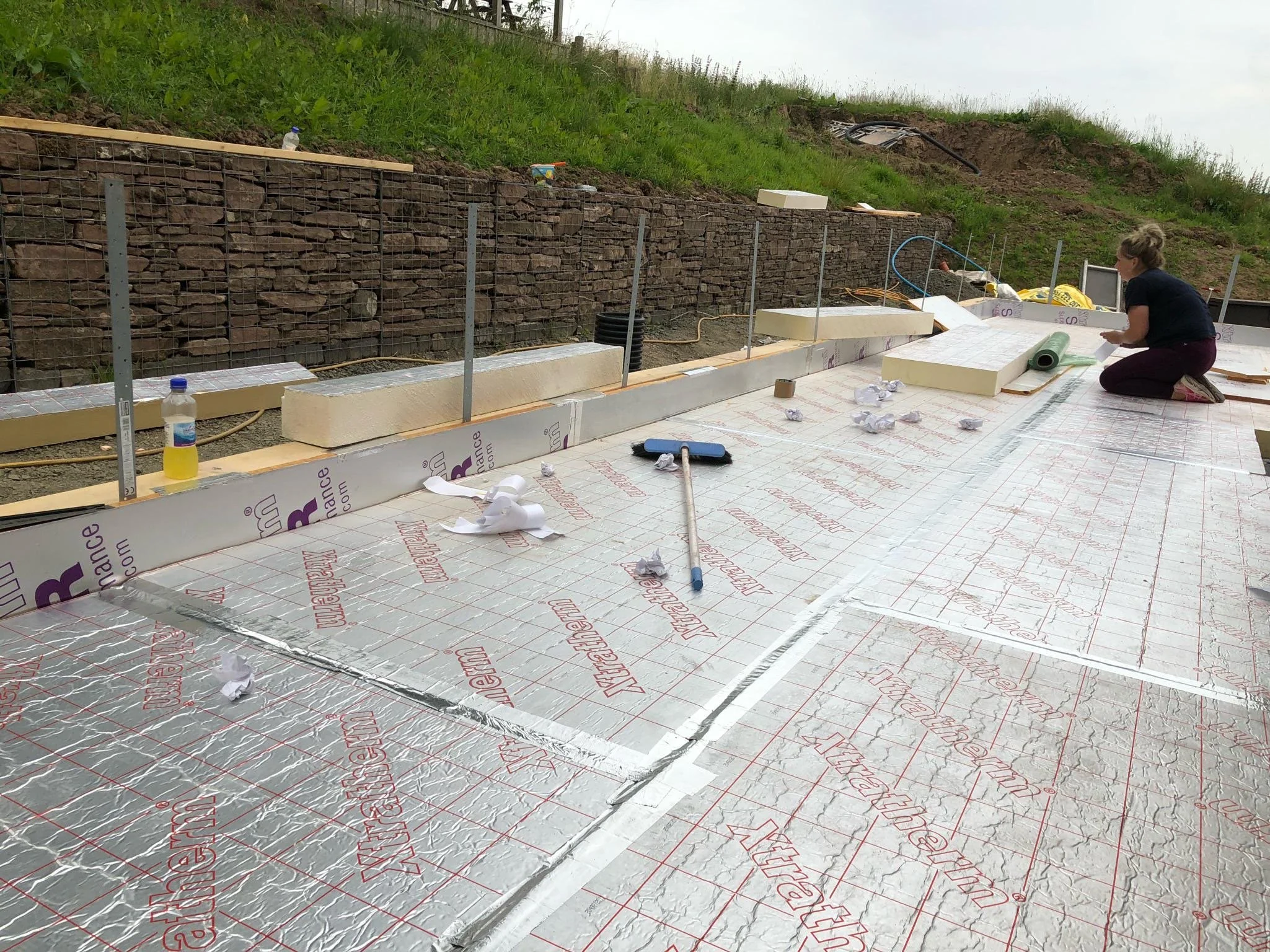 Construction worker kneeling on insulation foam at a building site with a stone retaining wall in the background and construction tools and materials scattered around.