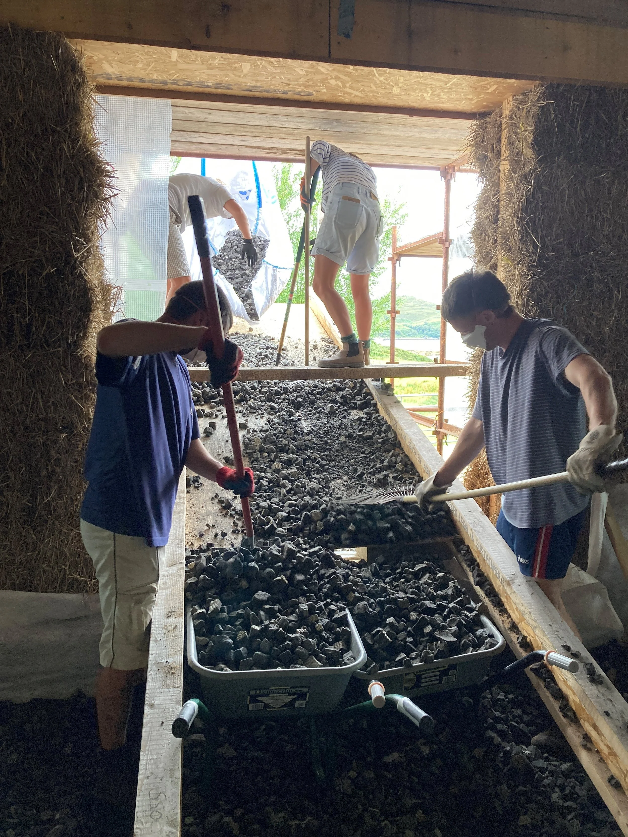 People working on a construction project inside a wooden structure, shoveling and moving rocks into a large container, with some of them wearing masks and gloves.