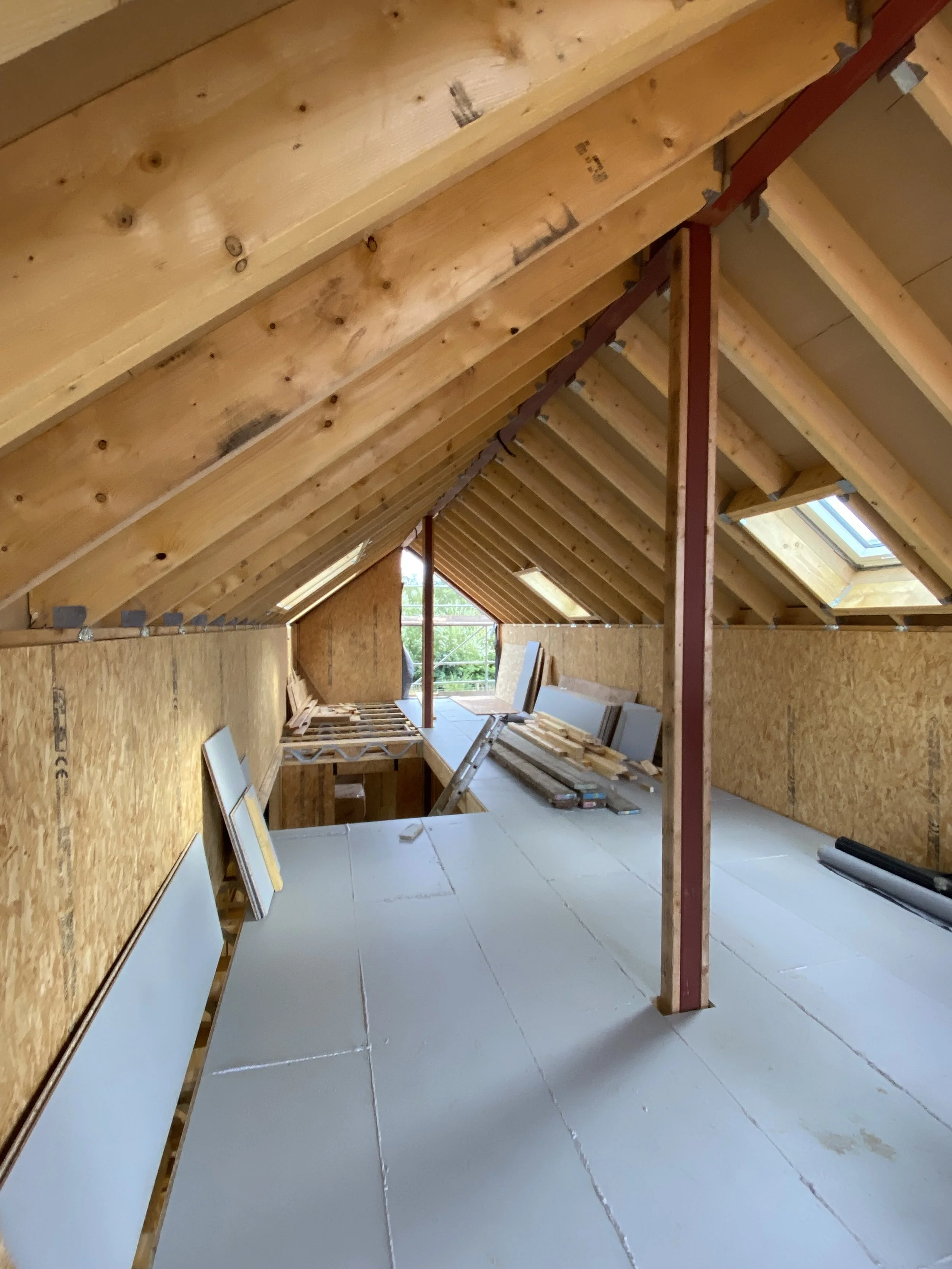 Interior of an attic or loft under construction, with wooden beams and insulation materials, skylight window, and construction tools and materials scattered around.