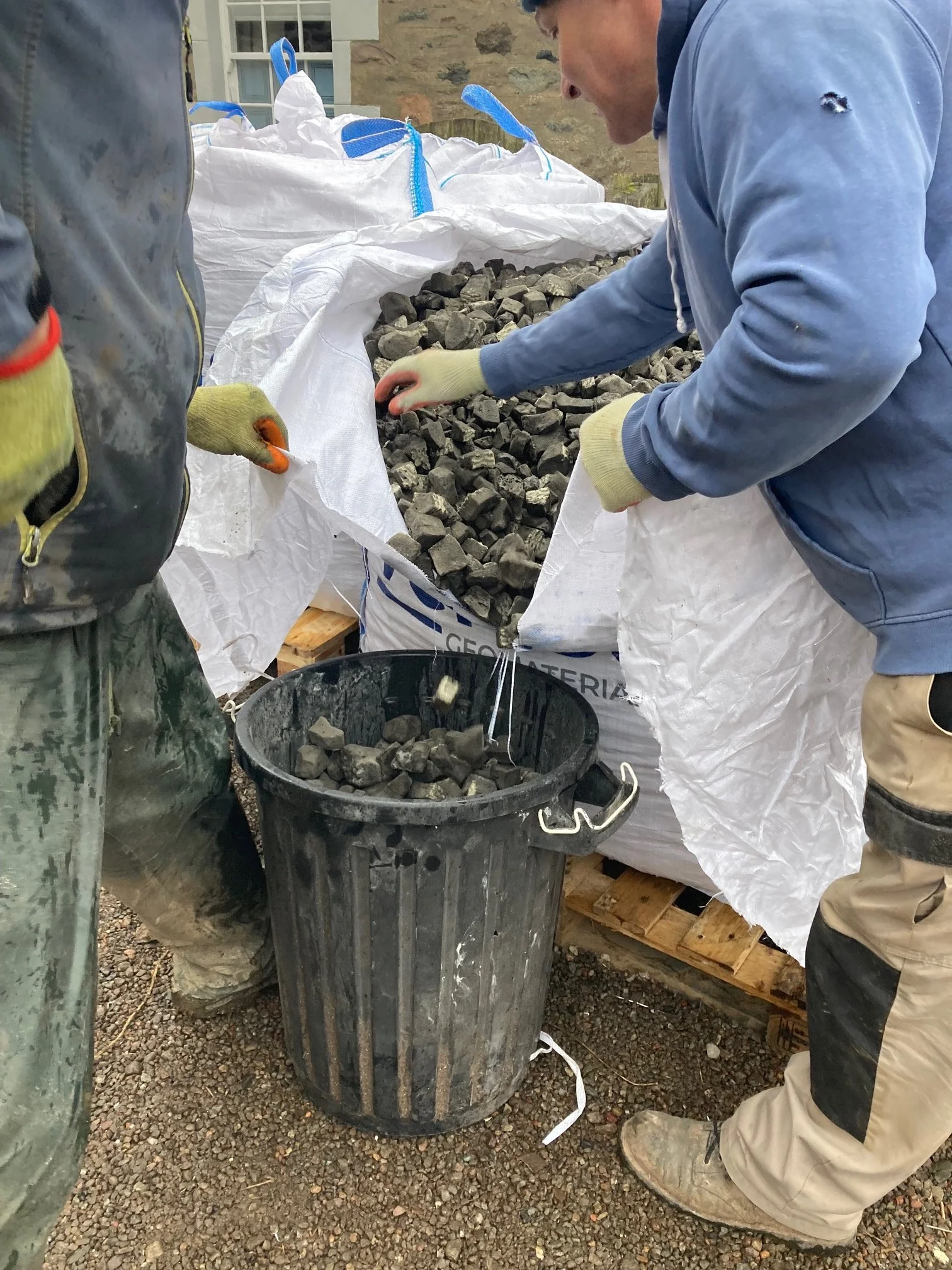 Two people wearing gloves are handling rough, dark stones from a large white sack that is on a wooden pallet outside. A black trash can is nearby.