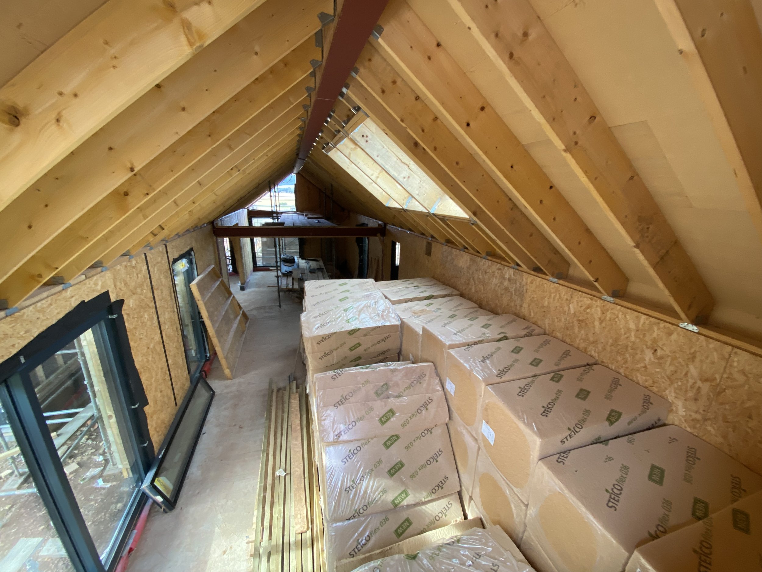 Interior of unfinished building with exposed wooden beams and insulation, construction materials stacked along the wall, and a large window showing an outdoor view.