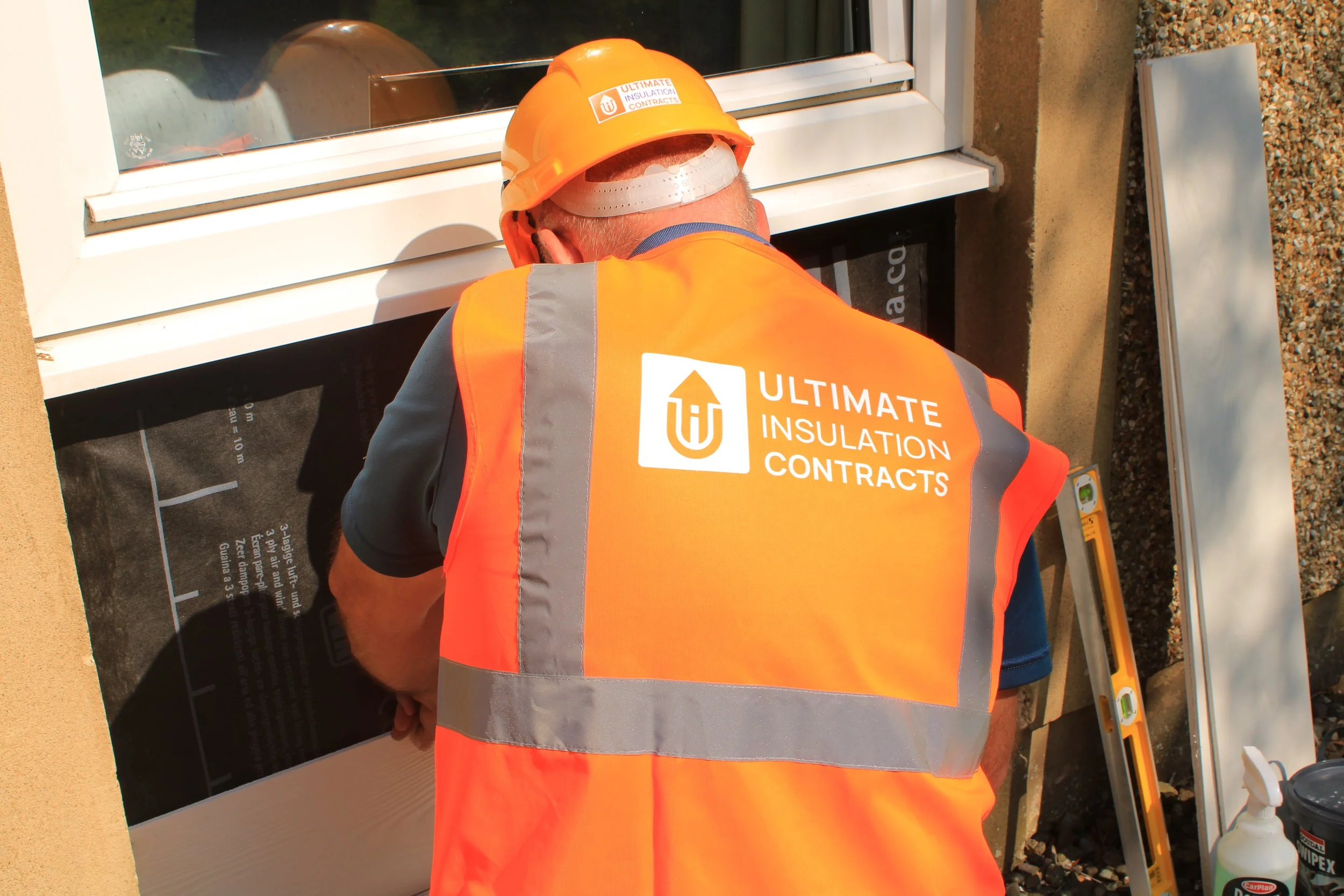 A construction worker wearing an orange safety vest and a yellow hard hat labeled 'Ultimate Insulation Contracts' is kneeling near a window opening, working on insulation or building materials.