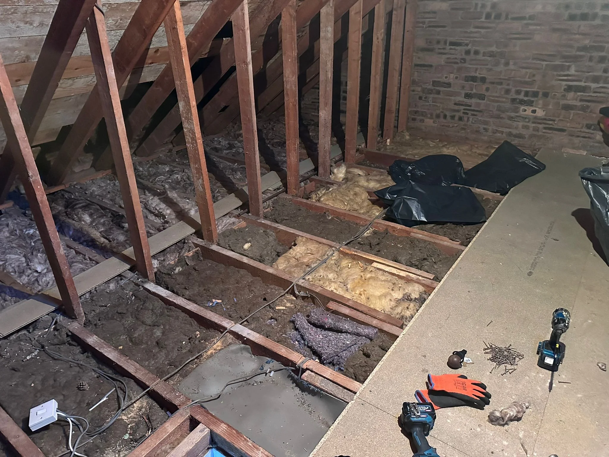View of a partially demolished area with exposed wooden floor joists, insulation, and a brick wall on the right. Tools such as a cordless drill, gloves, and various small equipment are scattered on the floor, indicating ongoing renovation work.
