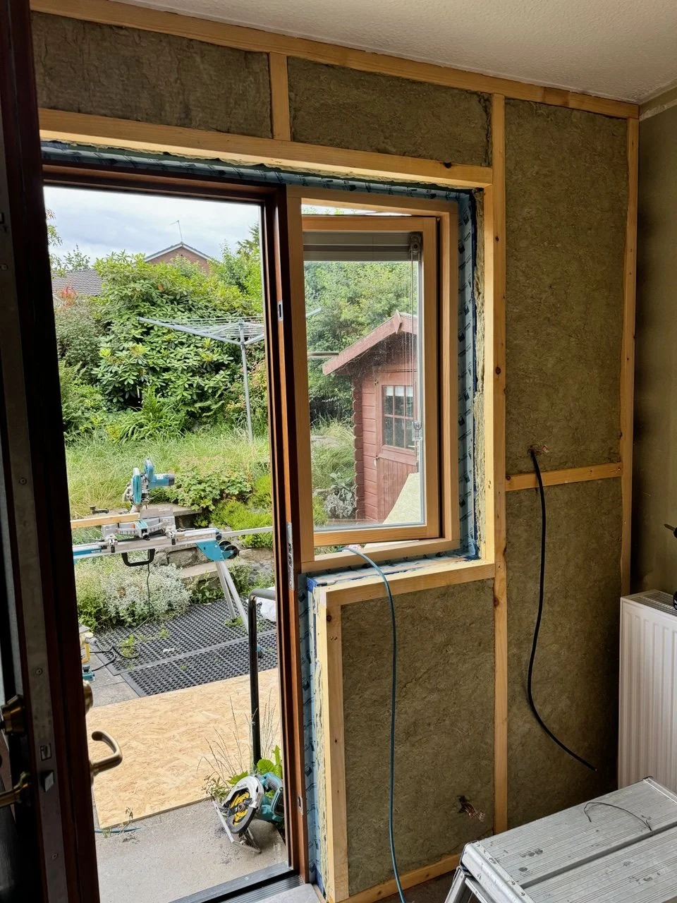 Inside a house under renovation showing a partially installed window with wooden framing and insulation. Outside, there is a garden with greenery, a wooden shed, and a workbench with power tools.
