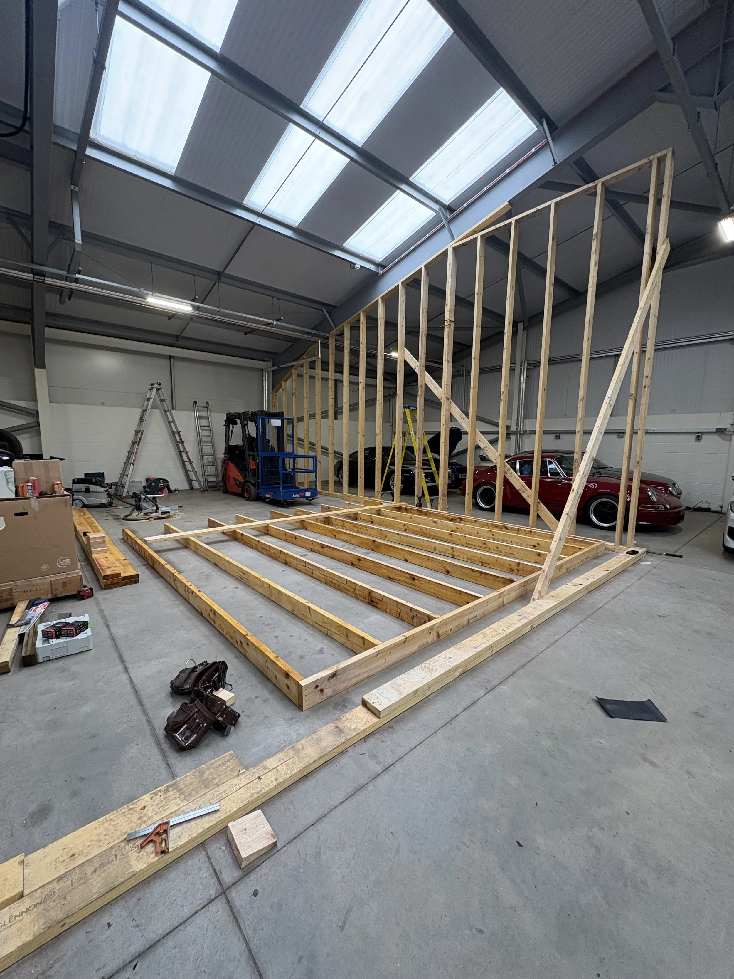 Construction site inside a warehouse with wooden framing for a wall, various tools, a ladder, and vintage cars parked inside.