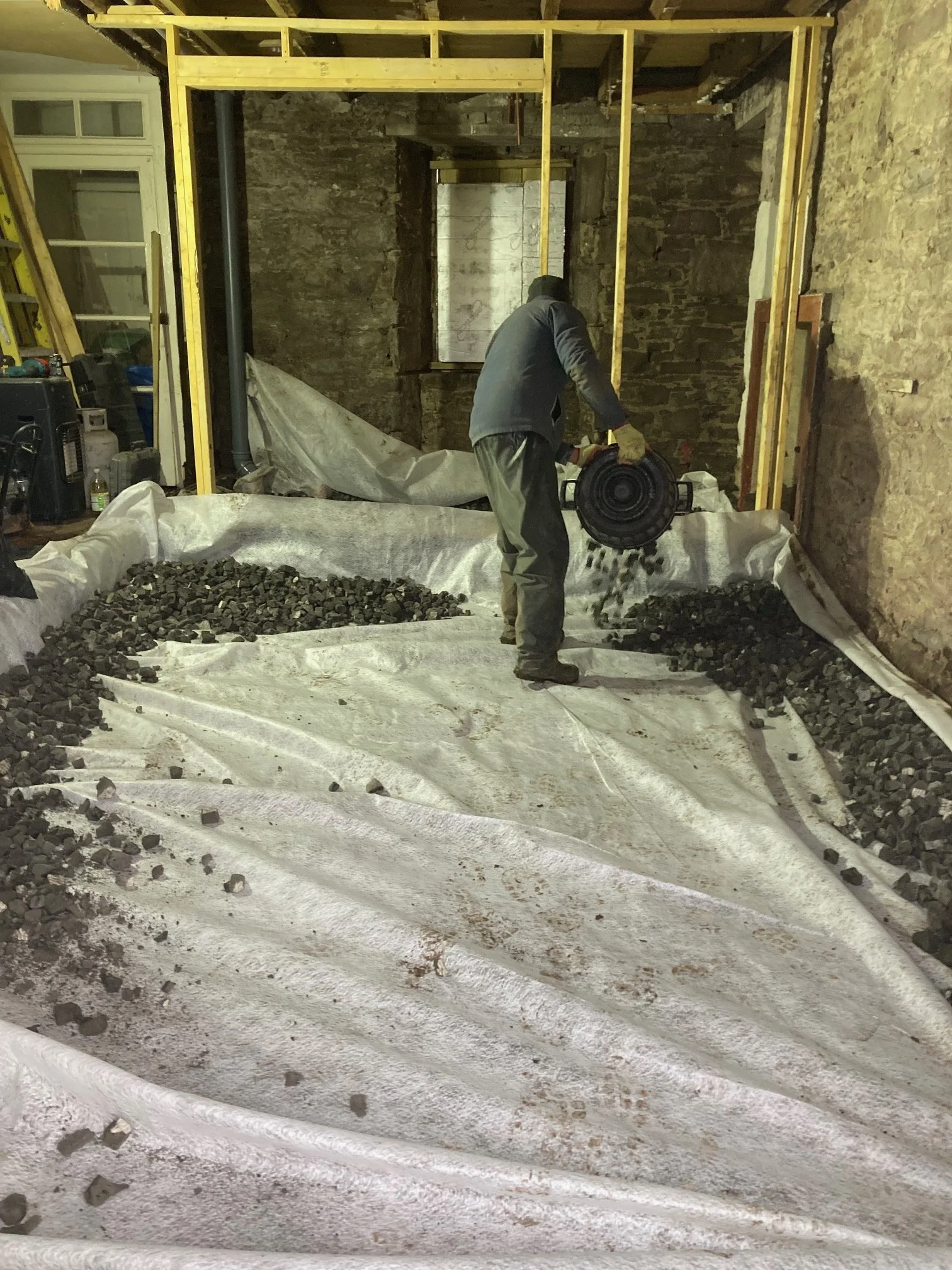 A person working on a construction project inside a room with exposed stone walls, using a power tool to work with rocks or gravel spread on a white tarp on the floor. The room has unfinished framing and a window opening.