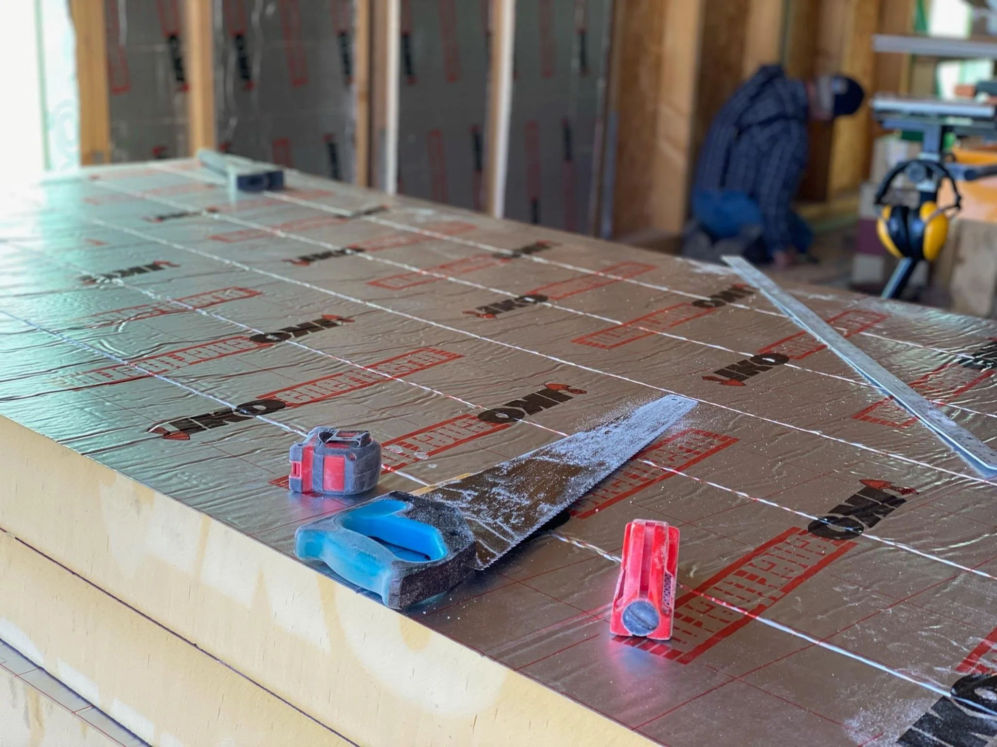 A construction site indoor with a partially finished wall and a work table covered with insulating foil material. Construction tools and safety equipment are visible around the area.