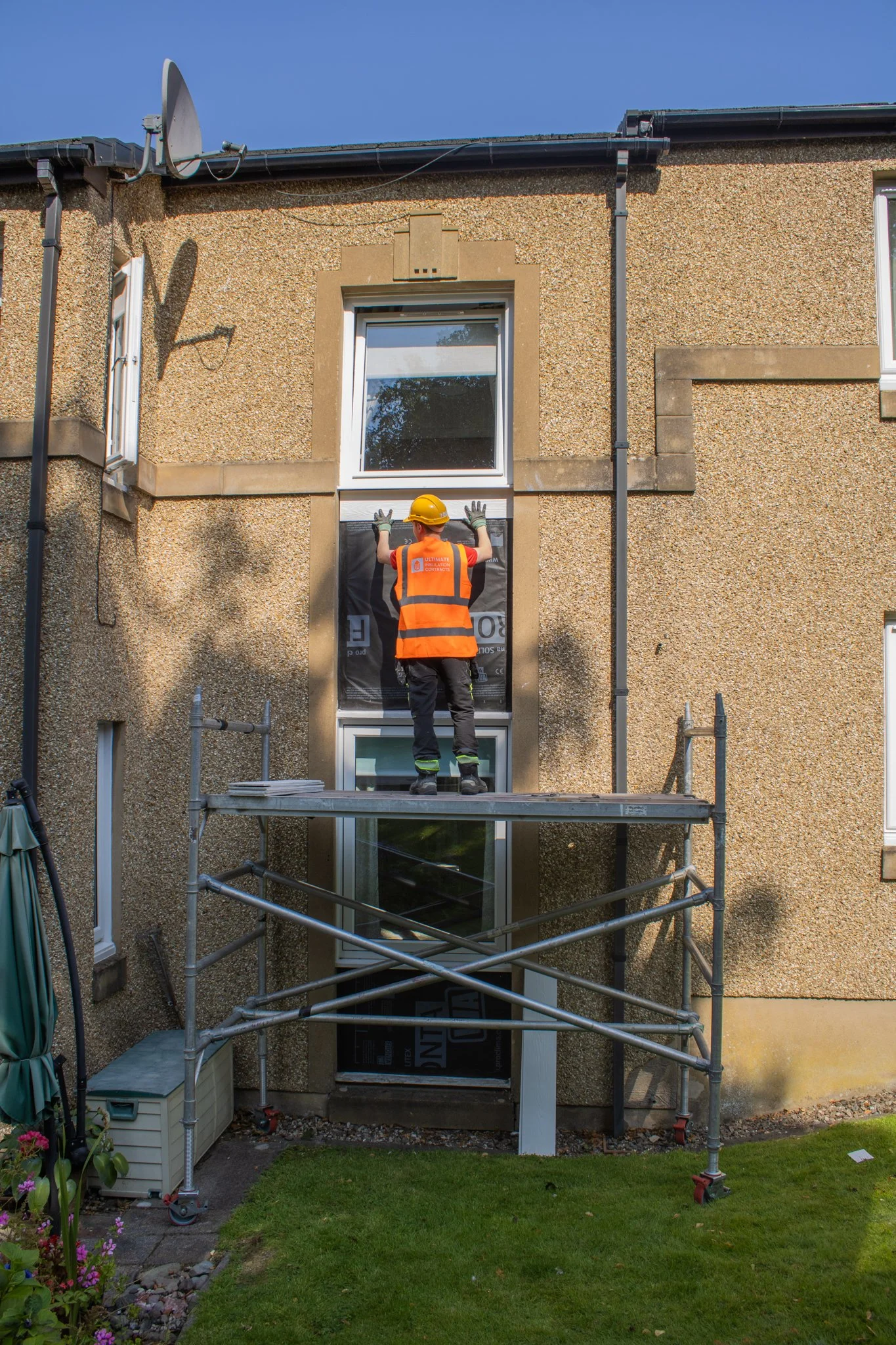 A construction worker in a yellow hard hat and orange safety vest is installing a new window on the second floor of a house. The house has a textured beige exterior wall with stone accents around the window frame and a scaffolding platform for access.