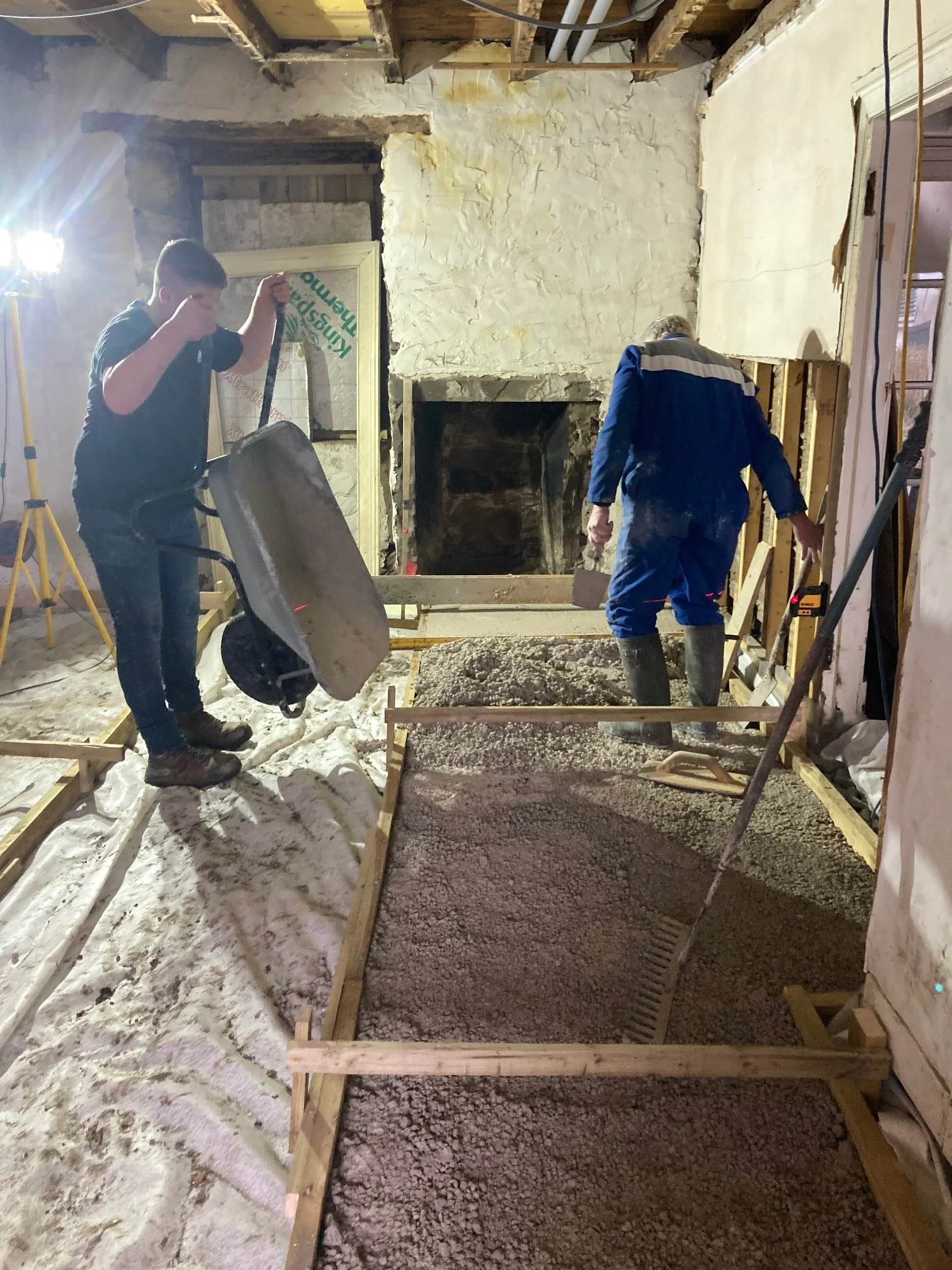Two workers are renovating a room, pouring and leveling concrete for a new floor. One person is using a wheelbarrow, and the other is spreading gravel or crushed stone. The room is in a state of construction with exposed wood beams, pipes, and unfinished walls.