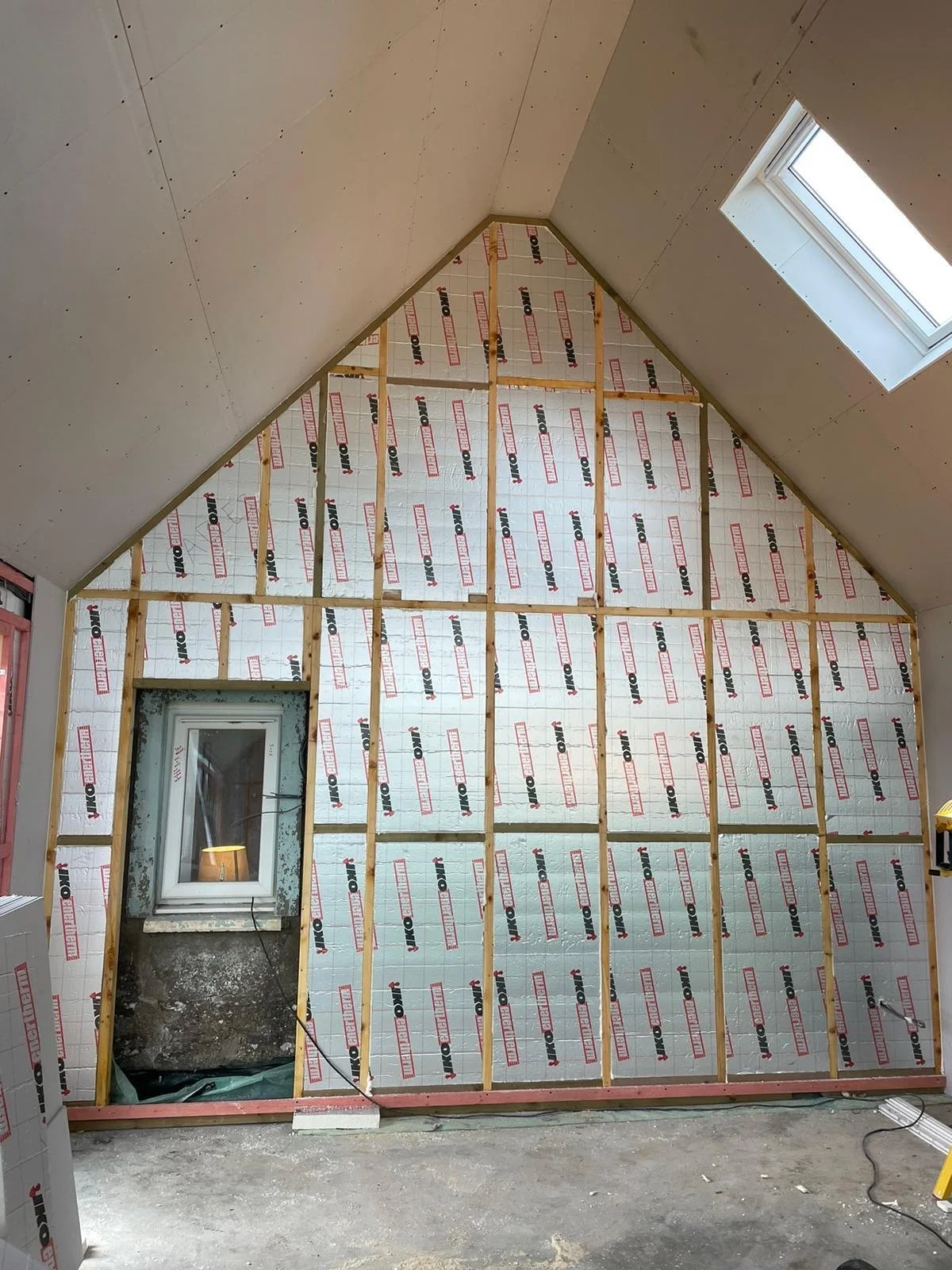 A construction site showing a partially built wall with insulation and wood framing inside a room with a sloped ceiling and a window.
