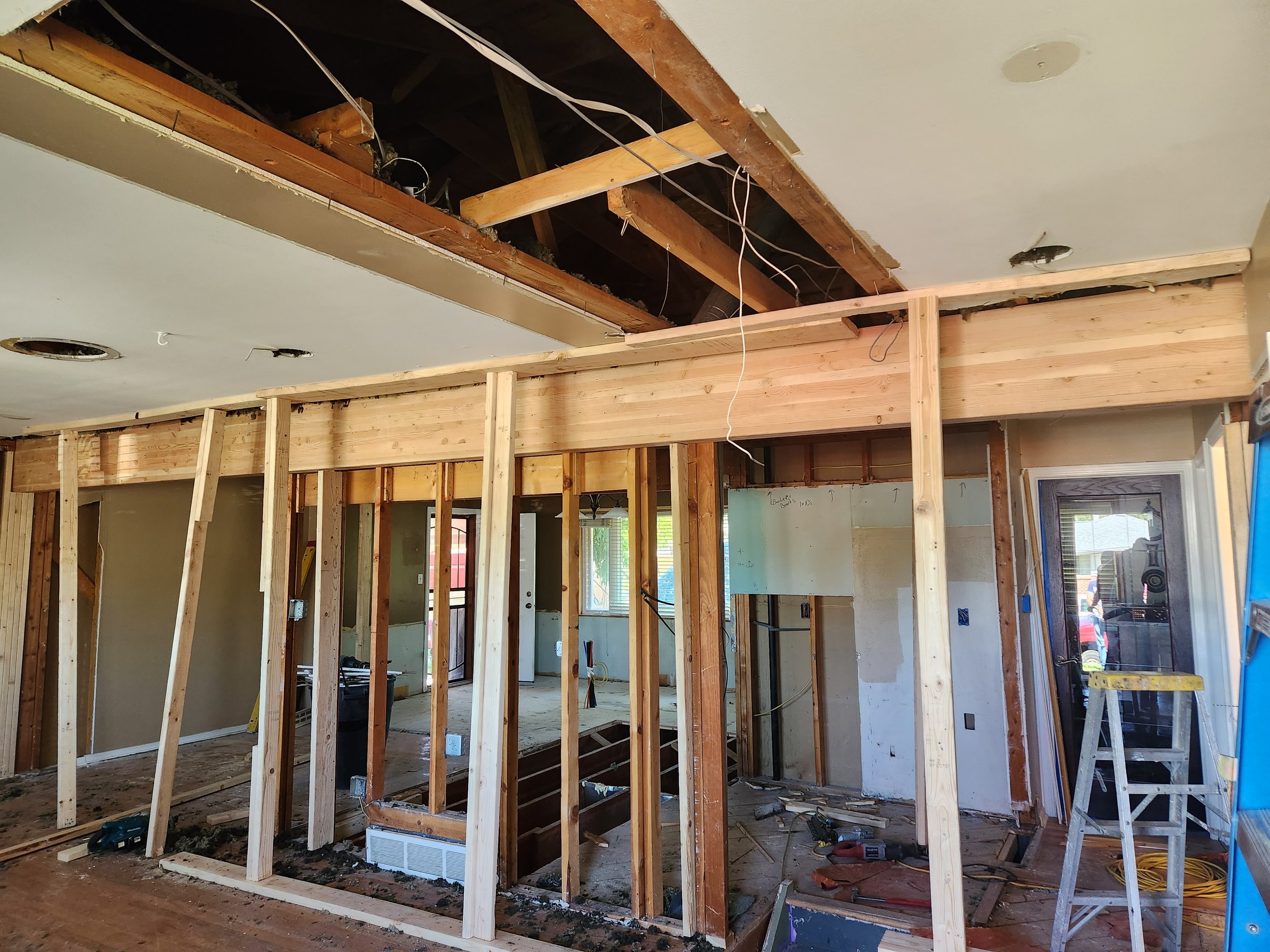 Interior of a house under renovation with exposed wooden framing, electrical wiring, and partially installed ceiling and wall panels.