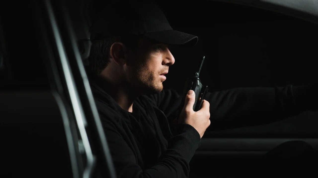 Ein Mann sitzt im Auto, schaut auf ein Funkgerät und trägt eine schwarze Mütze.