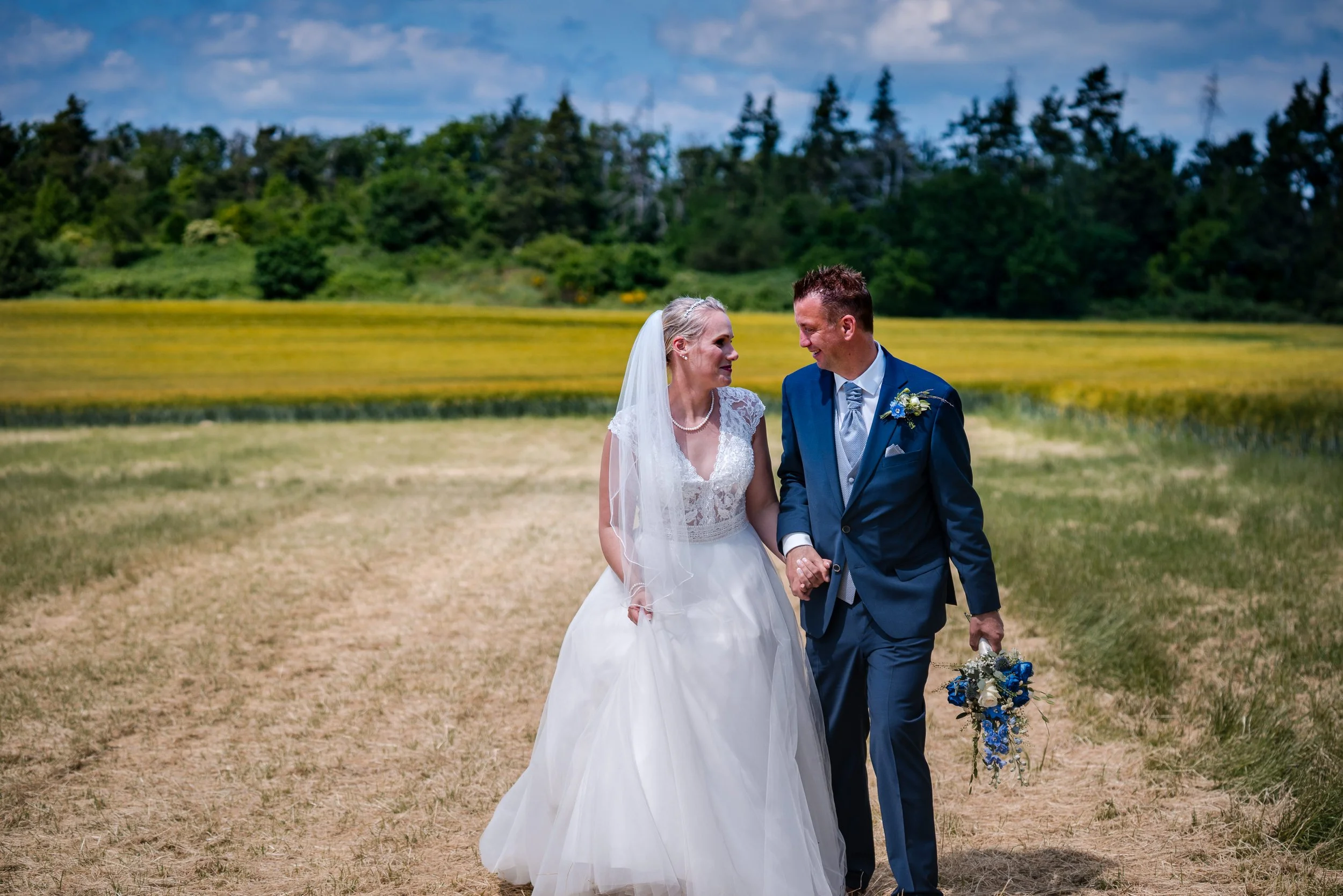 Ein Brautpaar läuft Hand in Hand auf einer Wiese bei einer Hochzeit. Der Himmel ist leicht bewölkt, im Hintergrund sind Bäume und grünes Land sichtbar.