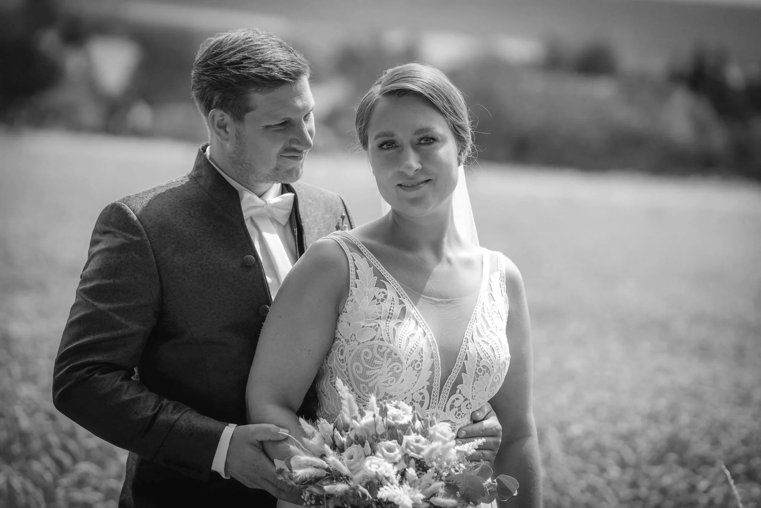 Ein Hochzeitsfoto eines Paares im Freien, schwarz-weiß Aufnahmen. Der Mann trägt einen traditionellen Anzug, die Frau ein schönes Kleid und hält einen Blumenstrauß. Sie stehen nah beieinander im Sonnenlicht, im Hintergrund eine offene Landschaft.