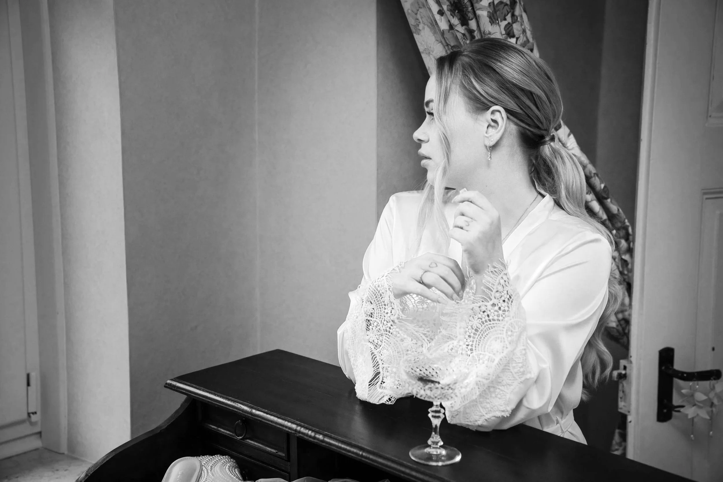 Eine Frau mit zartem Make-up und braunen, gelockten Haaren, die nach rechts schaut, trägt eine weiße, mit Spitze verzierte Robe. Sie steht neben einem schwarzen Tisch, der eine große, offene Schmuckschatulle und eine leere, halbliegende Weintraube au