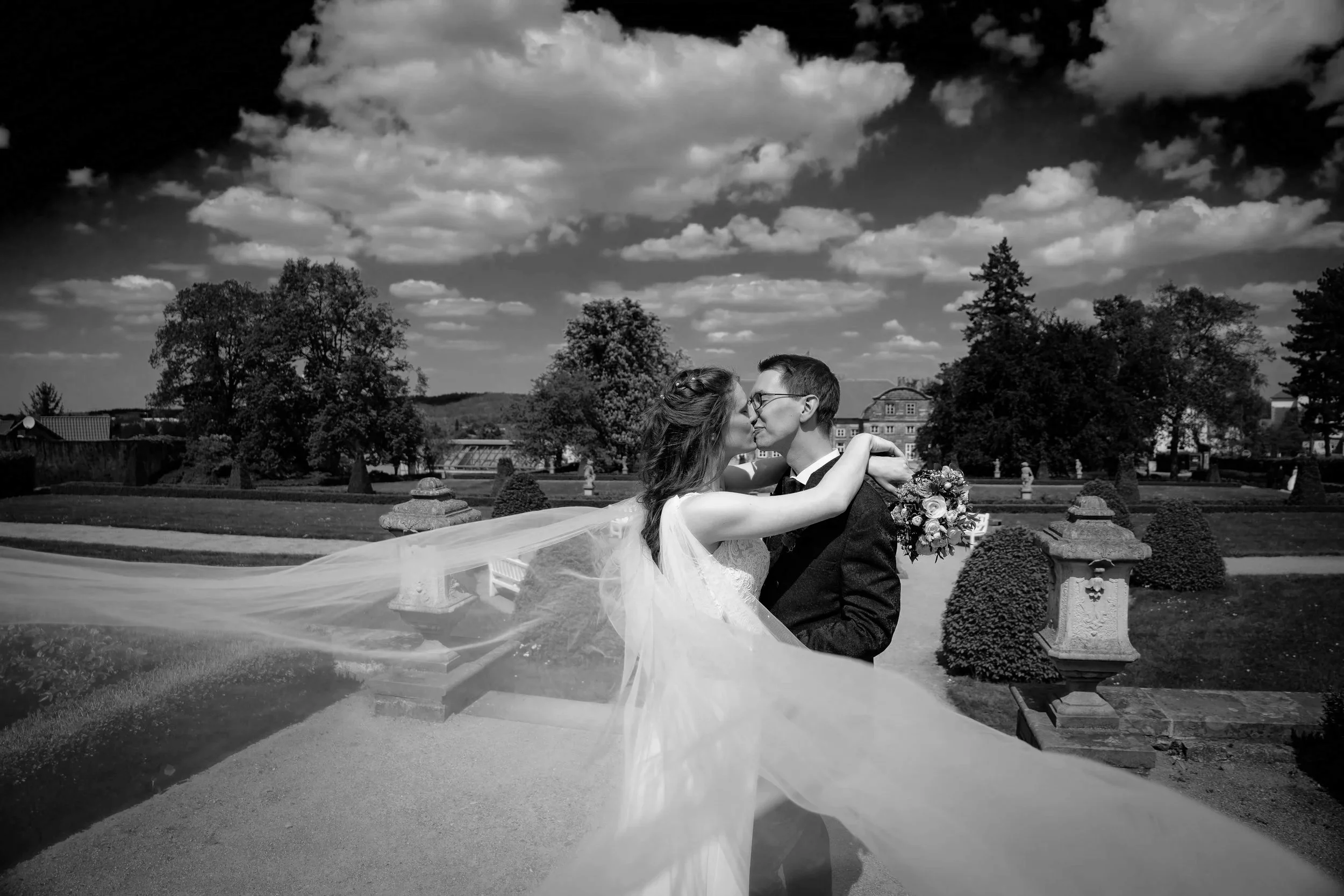 Ein Brautpaar küsst sich in einem Park bei schönem Wetter, umgeben von Bäumen und Skulpturen, im Schwarz-Weiß-Foto.
