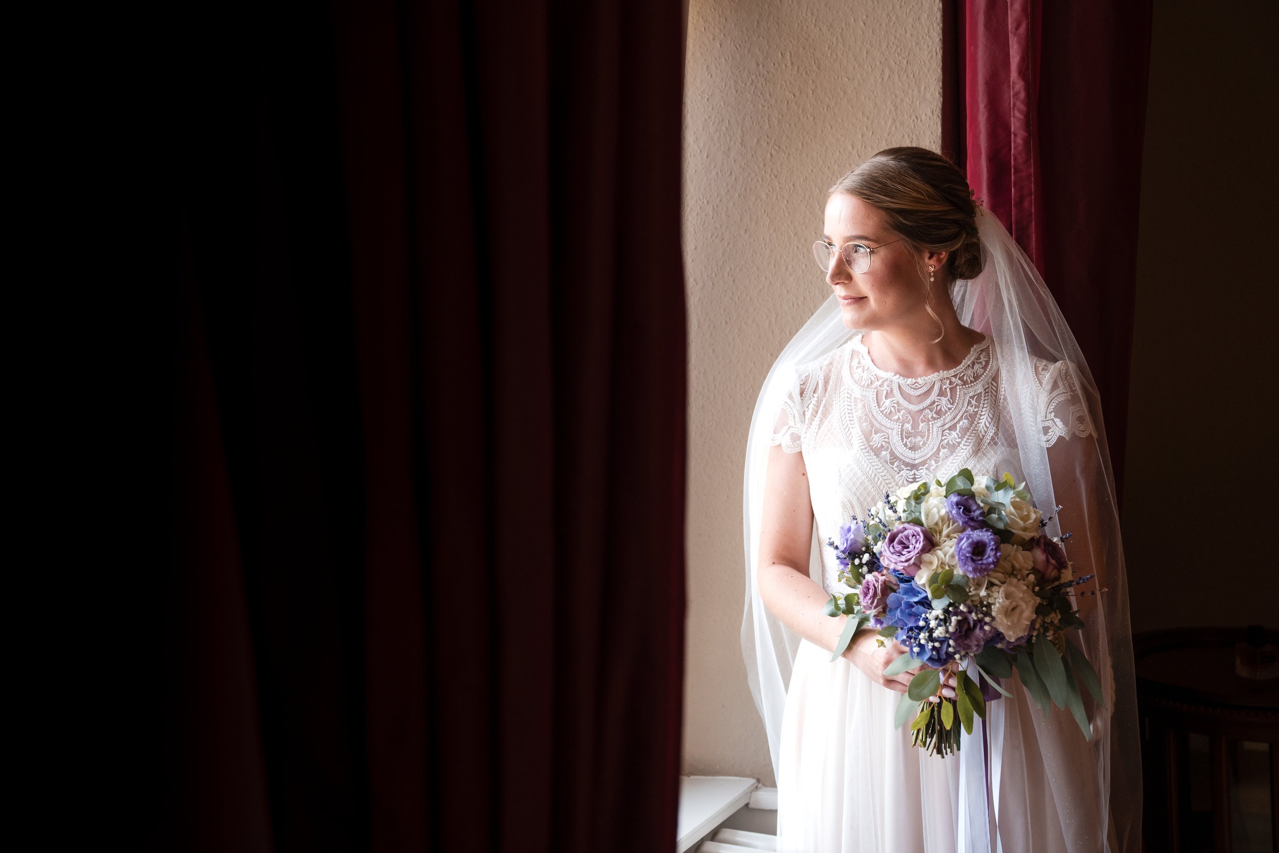 Braut mit Brille und Hochzeitskleid, Blick nach rechts, hält einen Blumenstrauß, steht vor einem Fenster mit roten Vorhängen.