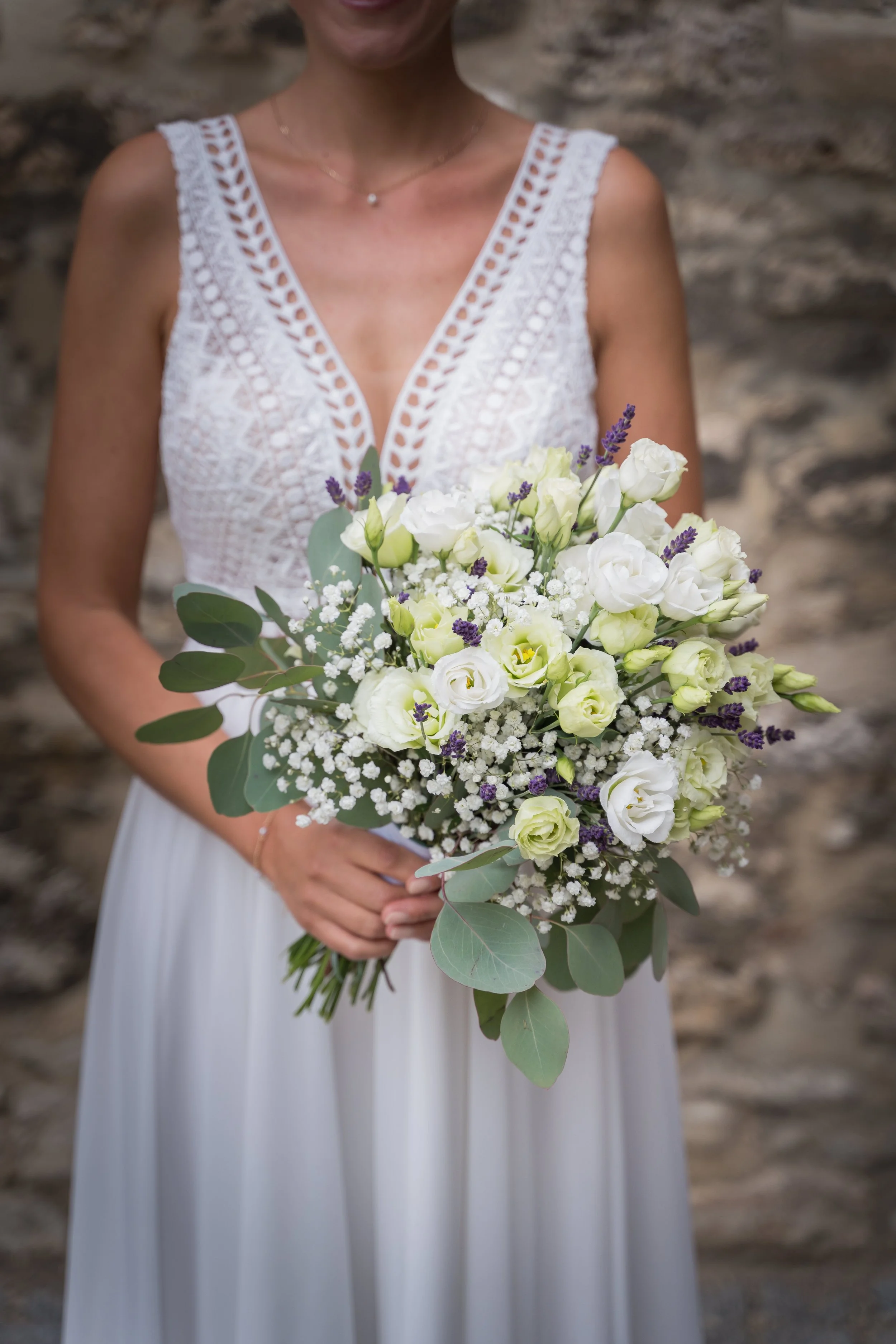 Frau in einem weißen Hochzeitskleid hält einen Blumenstrauß aus weißen und lila Blumen vor einem Steinmauer-Hintergrund.
