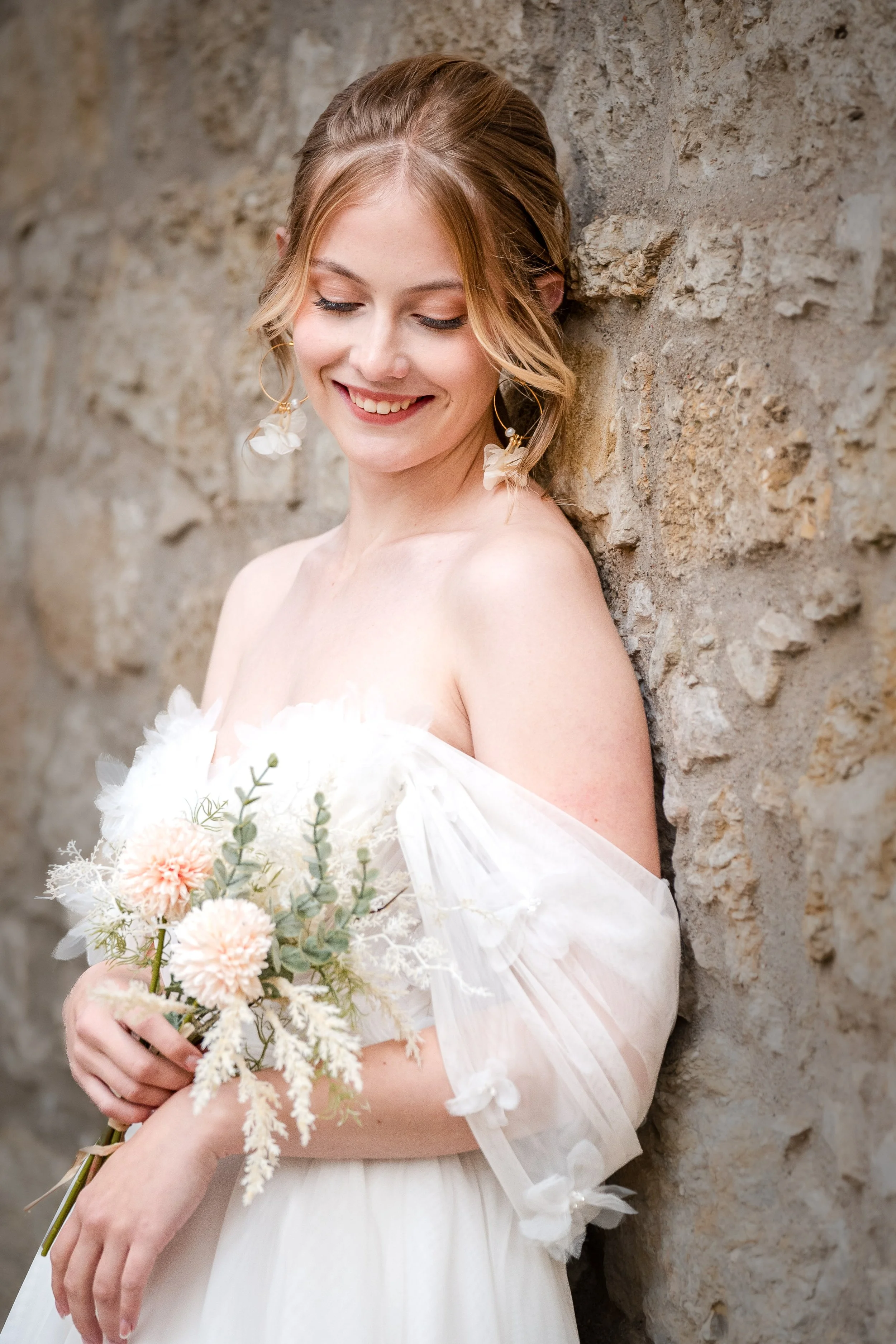 Ein Braut in einem Hochzeitskleid hält einen Blumenstrauß, lächelt und lehnt an einer Steinmauer.