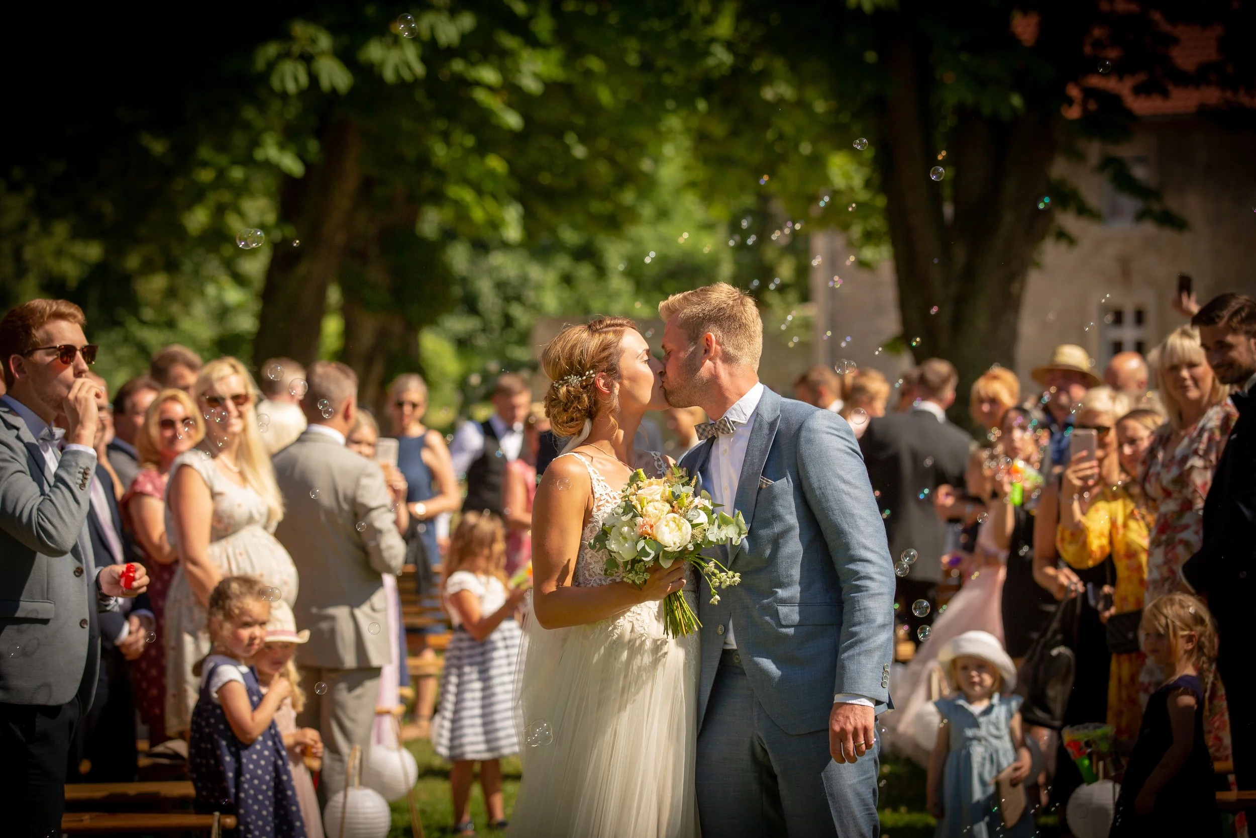 Ein frisch verheiratetes Paar küsst sich bei einer Hochzeit im Freien, umgeben von Freunden und Familie, mit Baum- und Gebäudeblenden im Hintergrund.