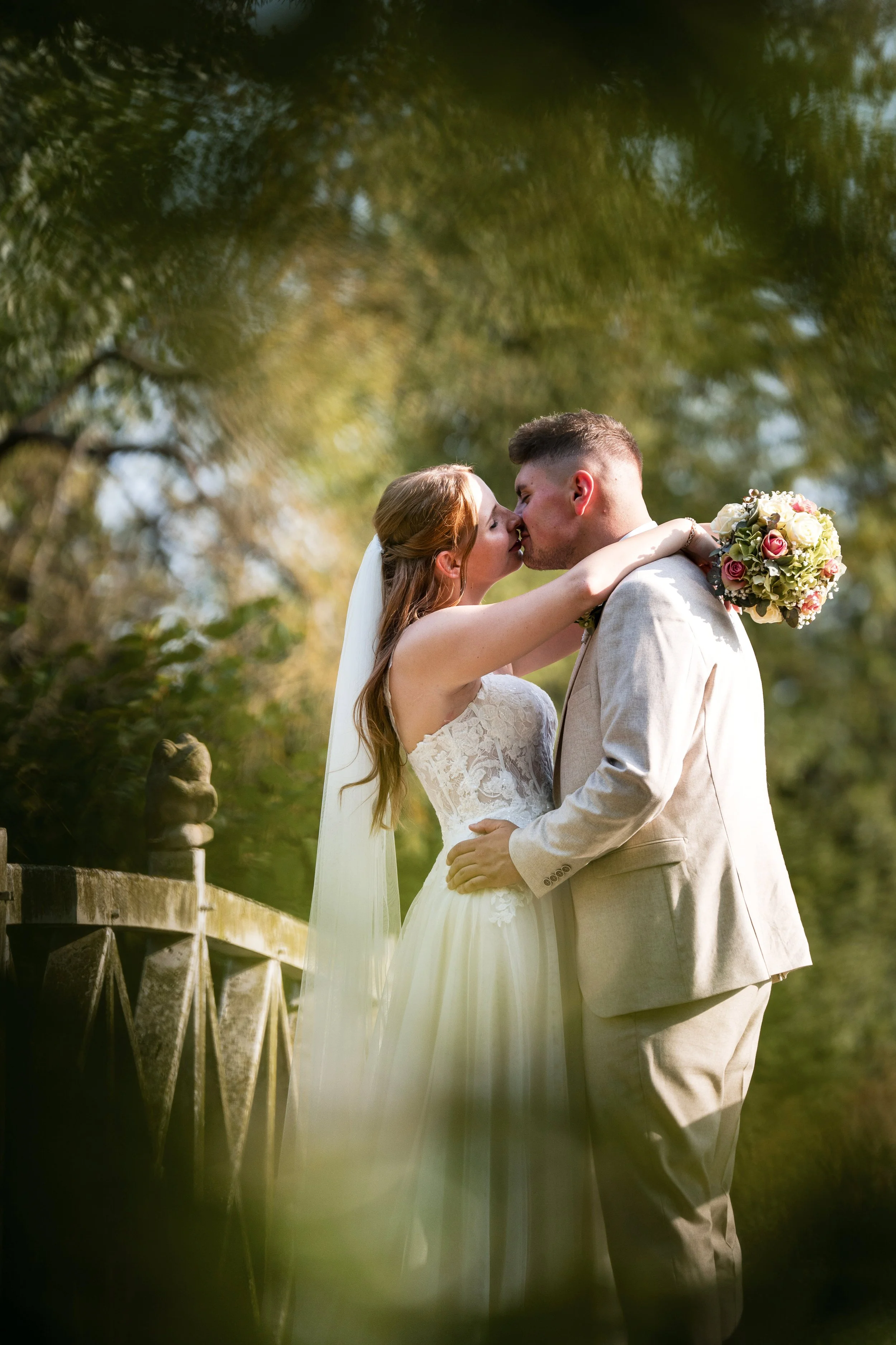 Ein Brautpaar bei einer romantischen Umarmung und Kuss im Freien an einem sonnigen Tag, die Braut trägt ein weißes Hochzeitskleid mit Spitze und einen Schleier, der Bräutigam einen hellen Anzug,  in ein tolle Location in Wolfenbüttel.