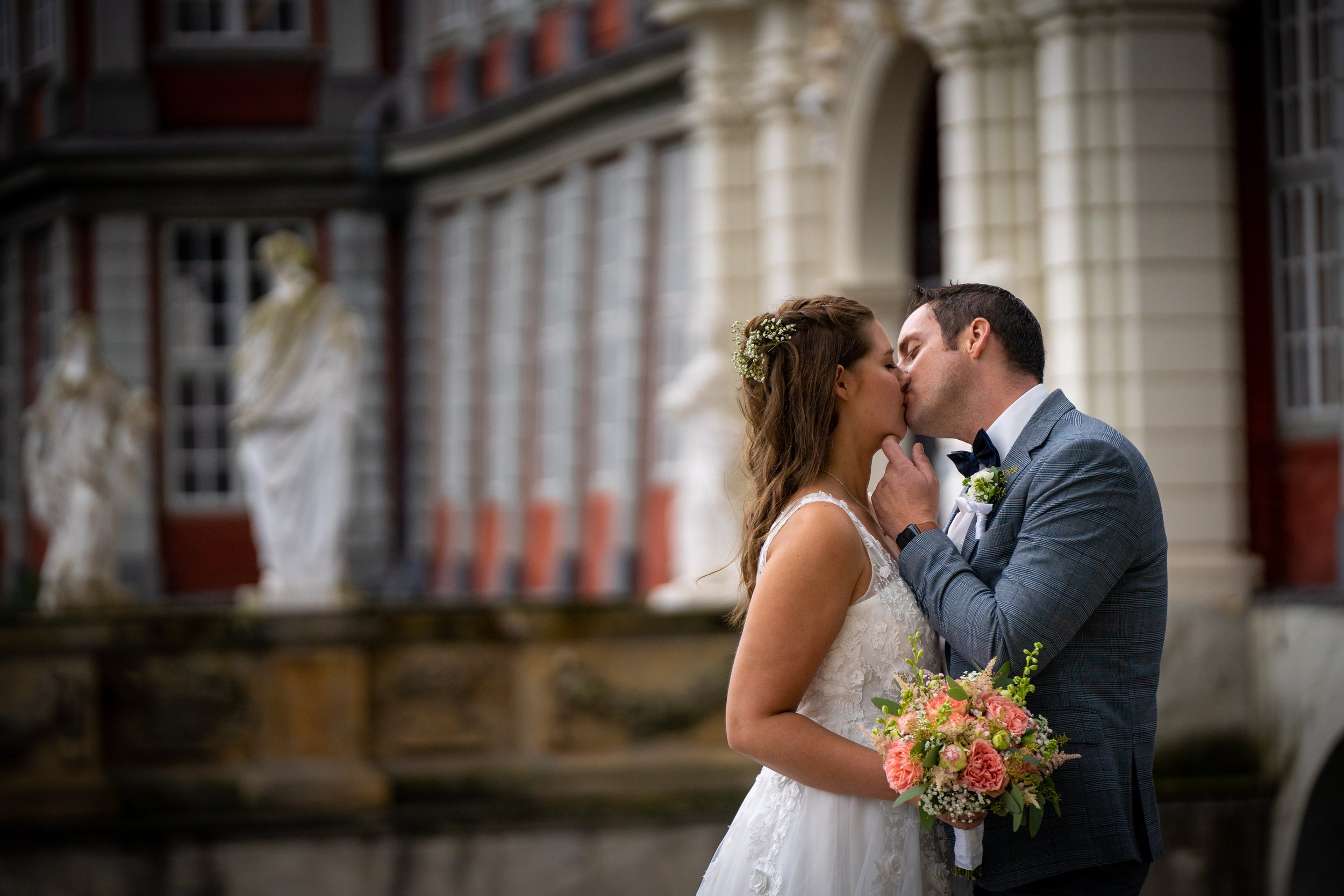 Ein Brautpaar bei einer Hochzeit, die Frau trägt ein weißes Hochzeitskleid und hält einen Blumenstrauß, der Mann trägt einen grauen Anzug. Sie küssen sich vor historischem Gebäude.